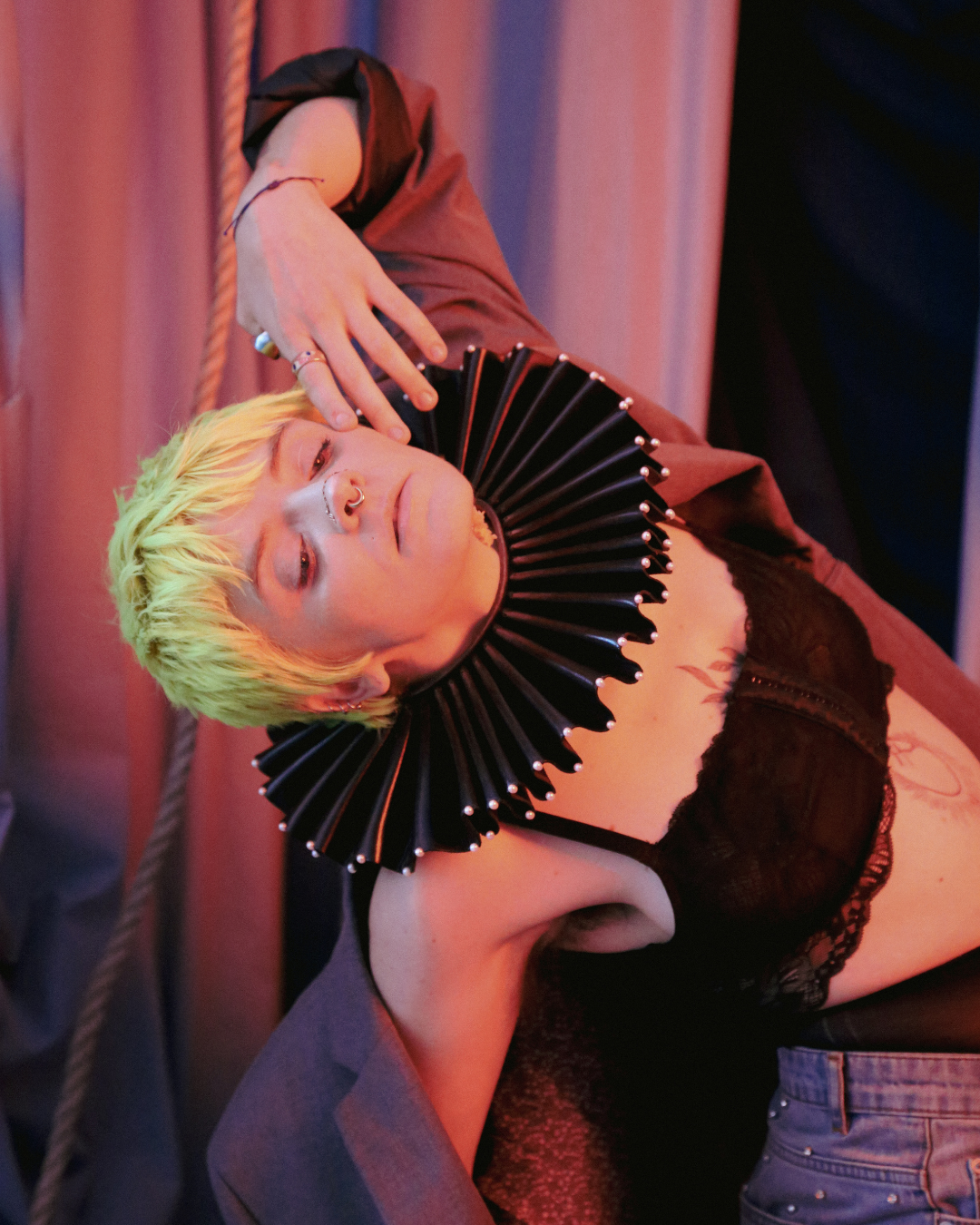 A lesbian poses with their hand pulling their mouth away and their back bent backwards. They have neon hair and a clown collar on. The photo has a pinkish overlay to it.