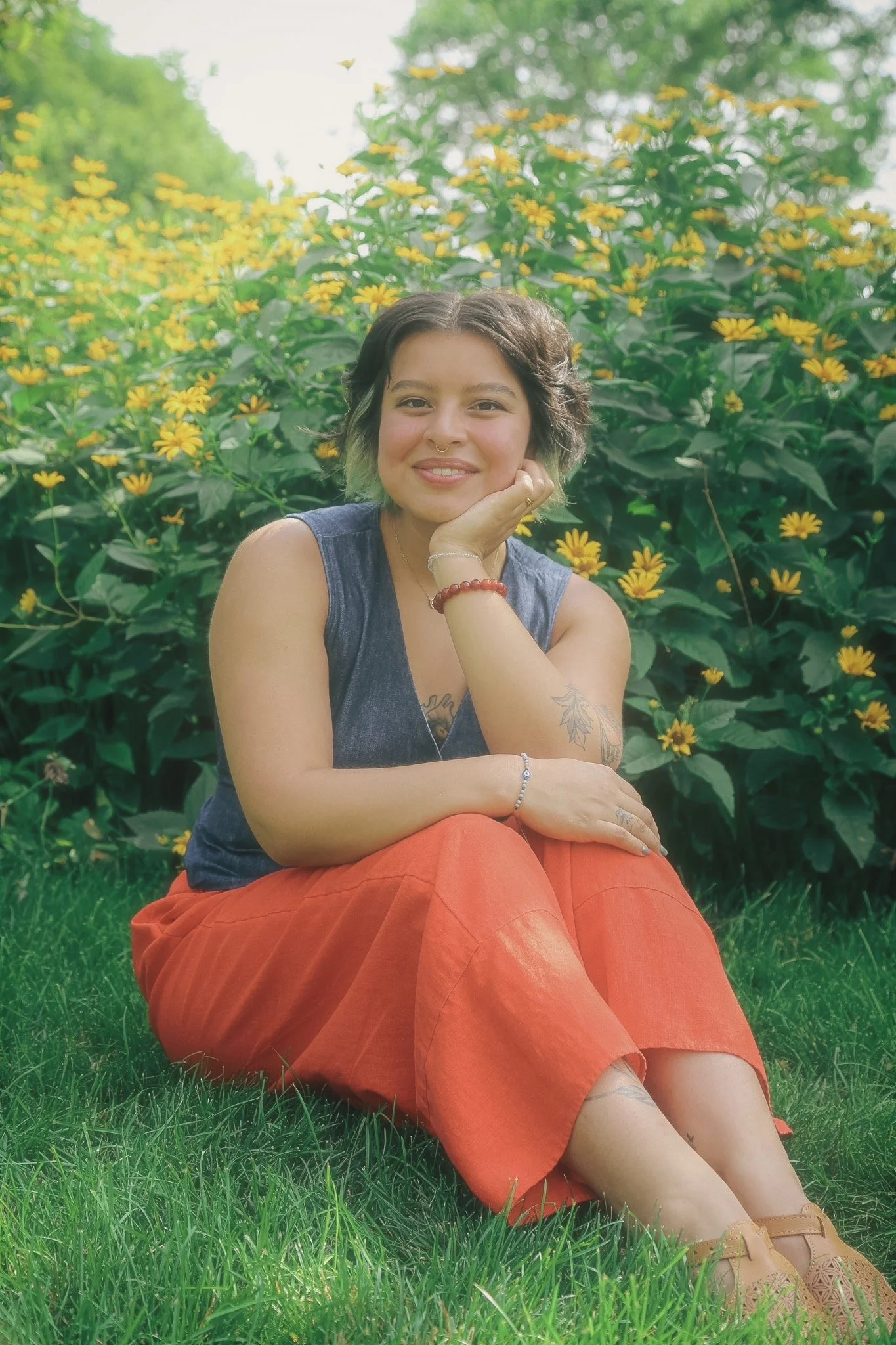 A person sits in a whimsical garden with green and yellow flowers. They have on a jean vest with orange pants. Their hair is brown and green. They are smiling into the camera. 