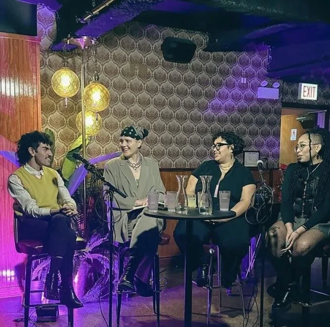 A group of queer photographers sit across from each other smiling. They are in a queer bar.  They are all sitting on stools and have mics in their hands. 