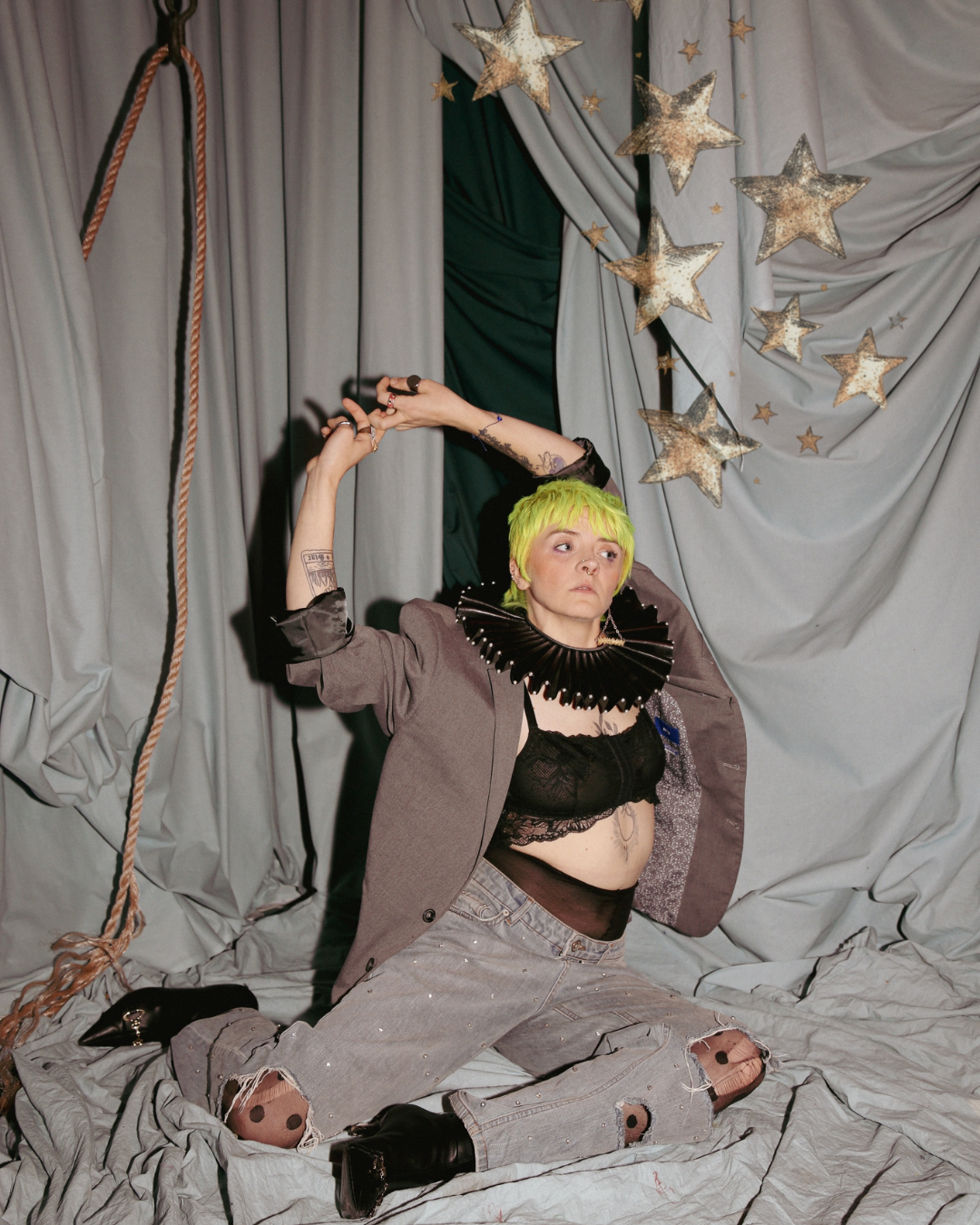 A lesbian poses on a grey sheet with a grey sheet background. They have their hands above their head and their legs contorted. Stars make their way above their head into the top of the photo.