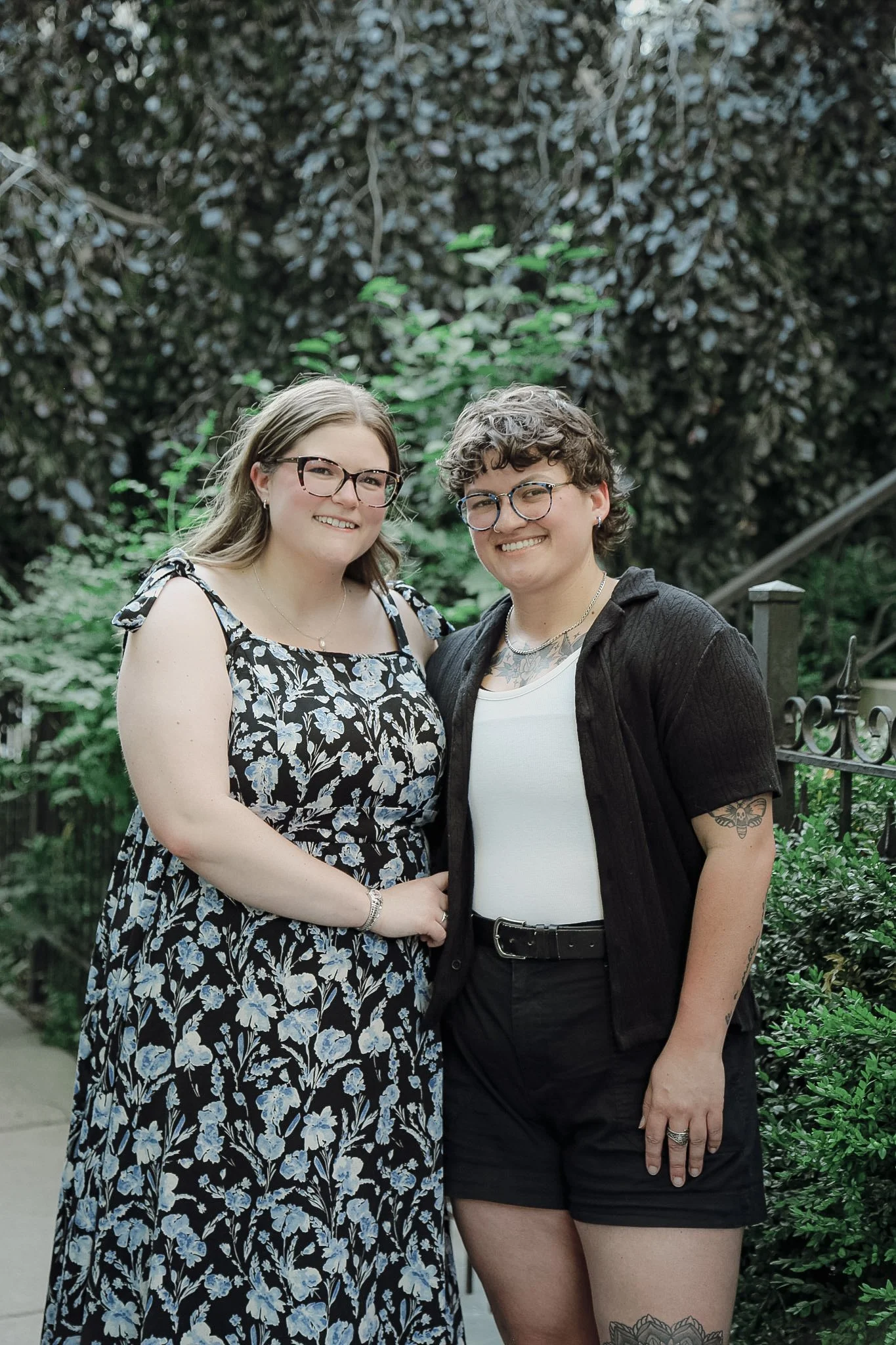Two queer people stand in front of a greenery covered wall. One with blonde hair and a blue & black dress. The other with short brown hair and a black top with black shorts. They are both smiling looking into the camera.