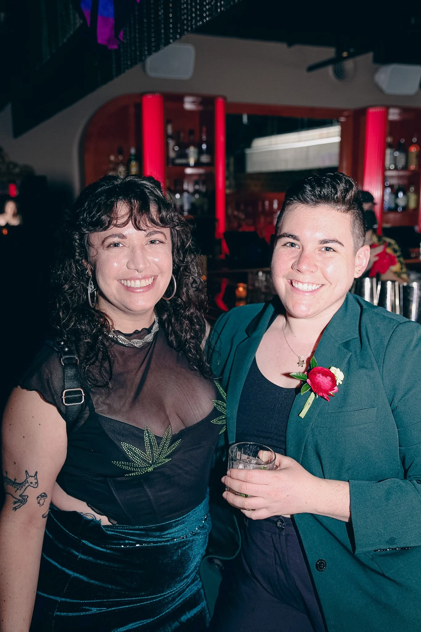 Two queer people stand side by side in a bar while wearing fancy outfits and smiling. One has curly brown hair and the other has shorter brown hair. They are color coordinating wearing green and black outfits.