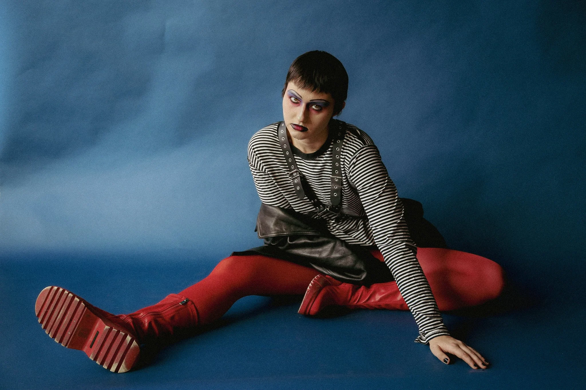 A queer person with blue, pink and black makeup sits seriously in front of a blue background. Wearing a leather jacket, striped black & white shirt, as well as red boots.