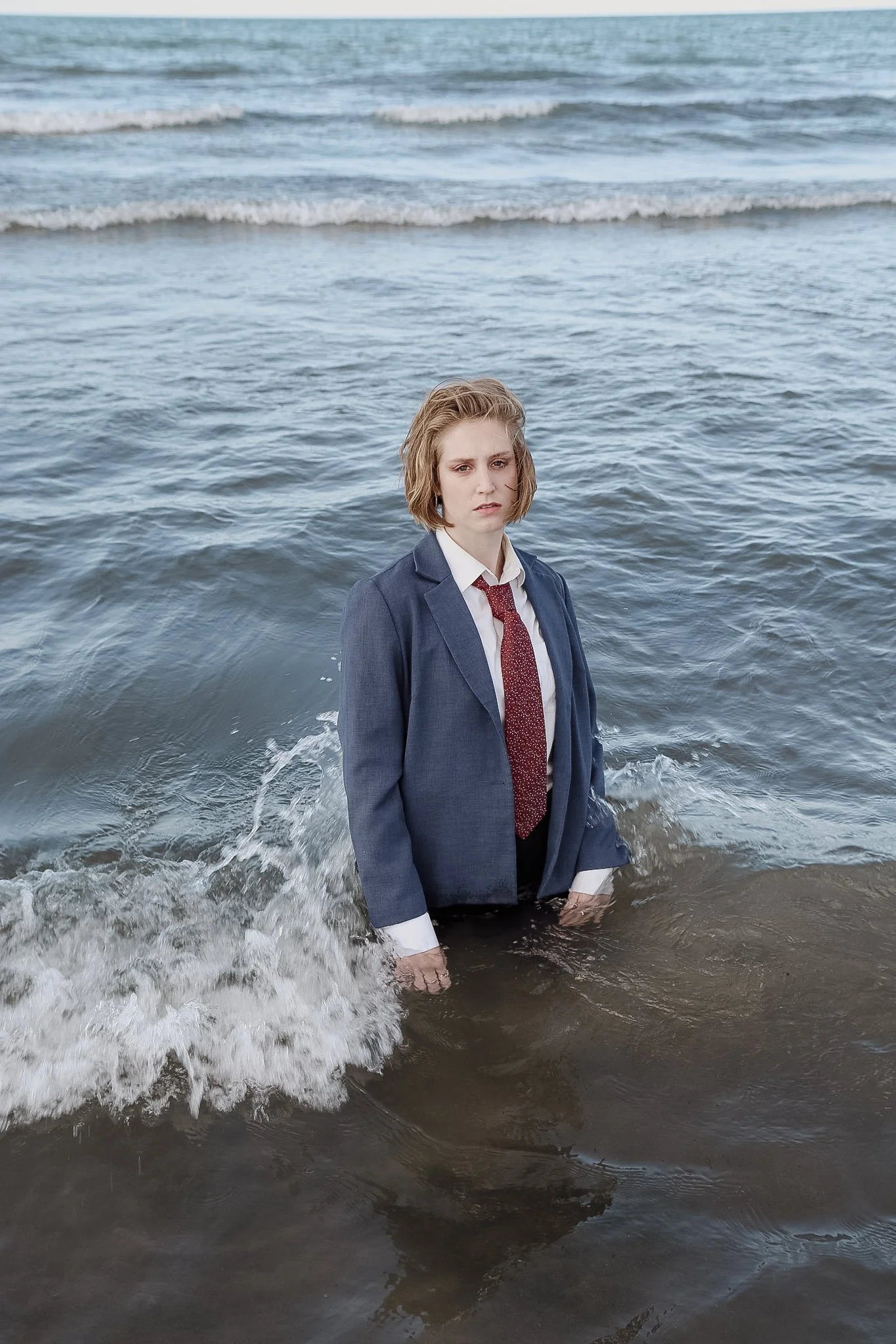 A queer person is standing waist-deep in water. The waves are splashing around them. The person has short blonde hair, a blue tux jacket, a white shirt, and a red tie. They are looking seriously into the camera.