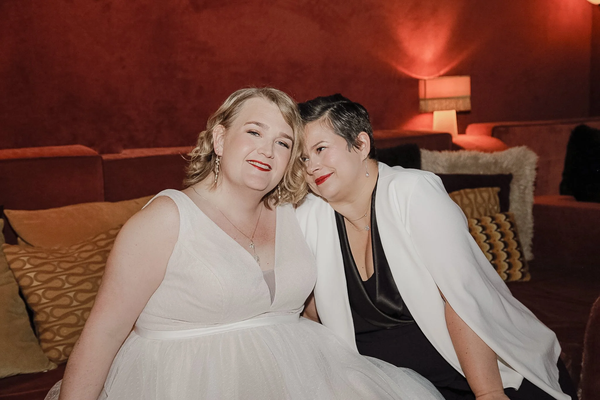 Two queer people who have just gotten married sit on a velvet orange couch. One person smiles at their partner wearing a black and white pantsuit. The other is wearing a white dress and is smiling into the camera.