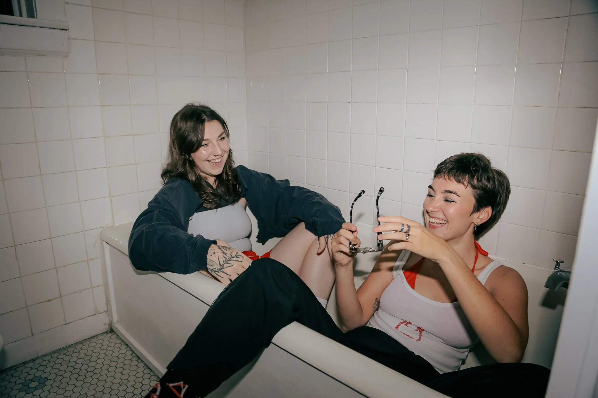Two queer people sit fully clothed in a bathtub laughing. Both people are wearing white, red and black outfits. One person is holding sunglasses in their hands.