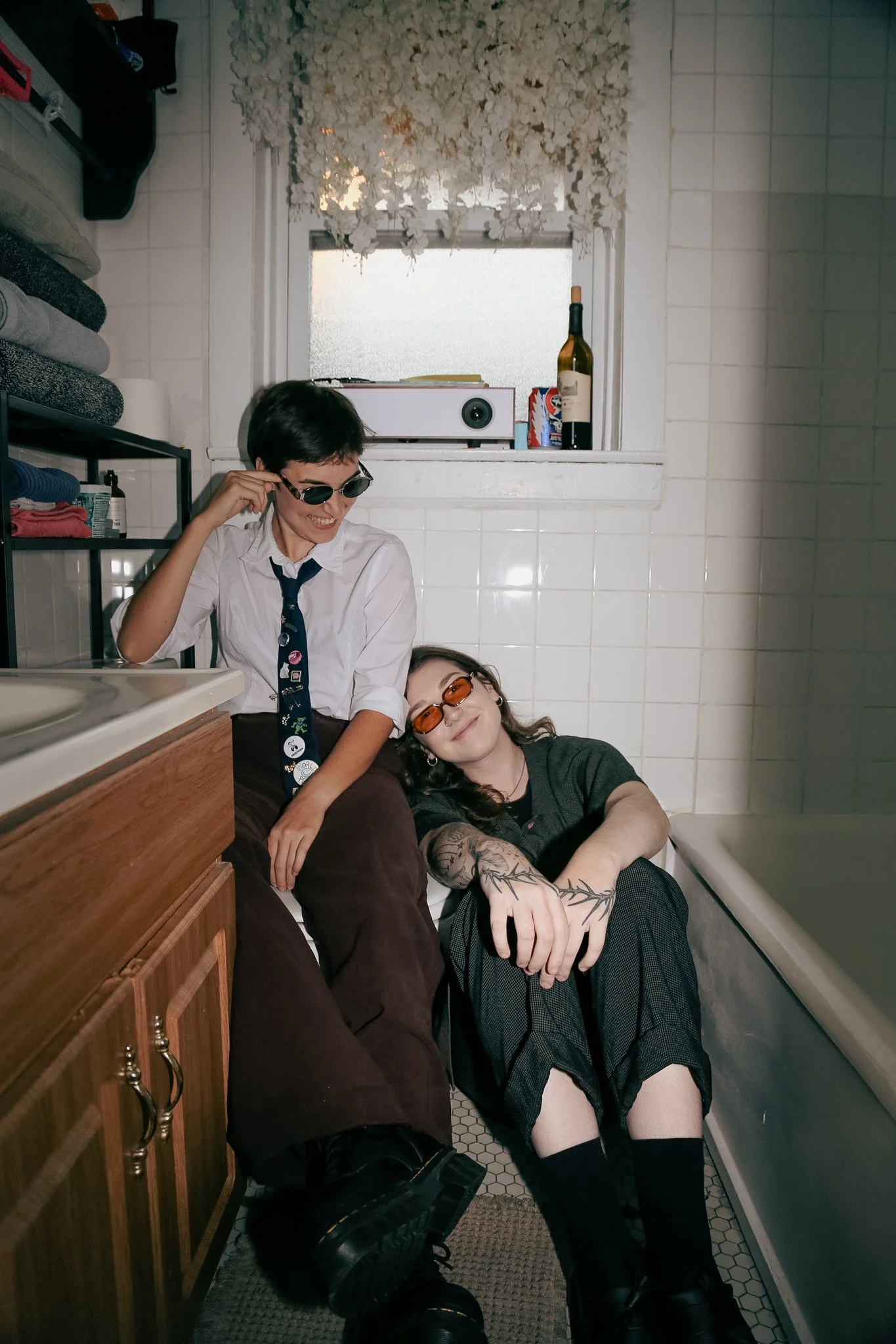 Two queer people sit side by side in a bathroom. One is laughing sitting on the toilet looking down at the other who is on the ground leaning their head on the other person. They have on business casual clothing and sunglasses.