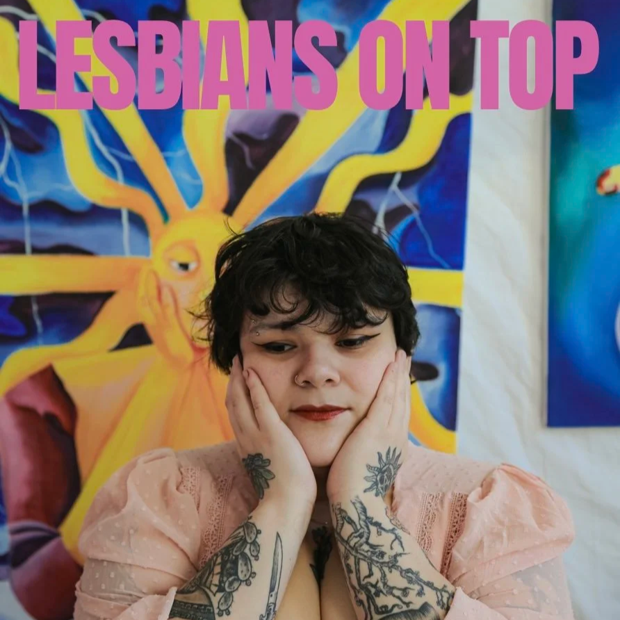 The photo highlights the photo series called "Lesbians On Top". A person with short, black curly hair sits in front of a painting with their hands on their face looking down. The top of the photo reads “Lesbians On Top”