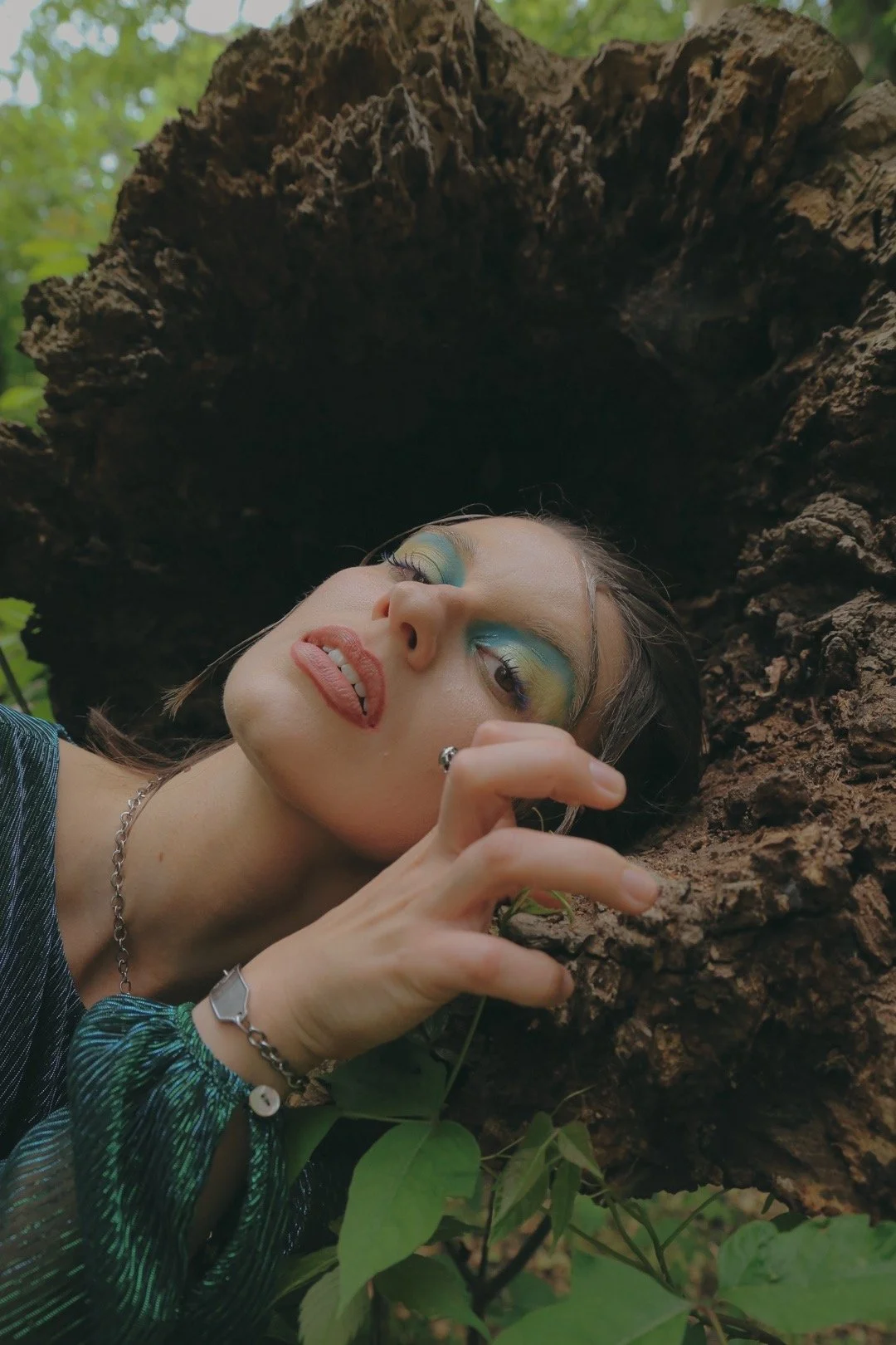 A person is leaning against a piece of wood in the forest with their hand clenched on a stump and their facial expression is serious. They have dark brown hair with green & yellow eye makeup that matches their green top. Instagram @marytrespass