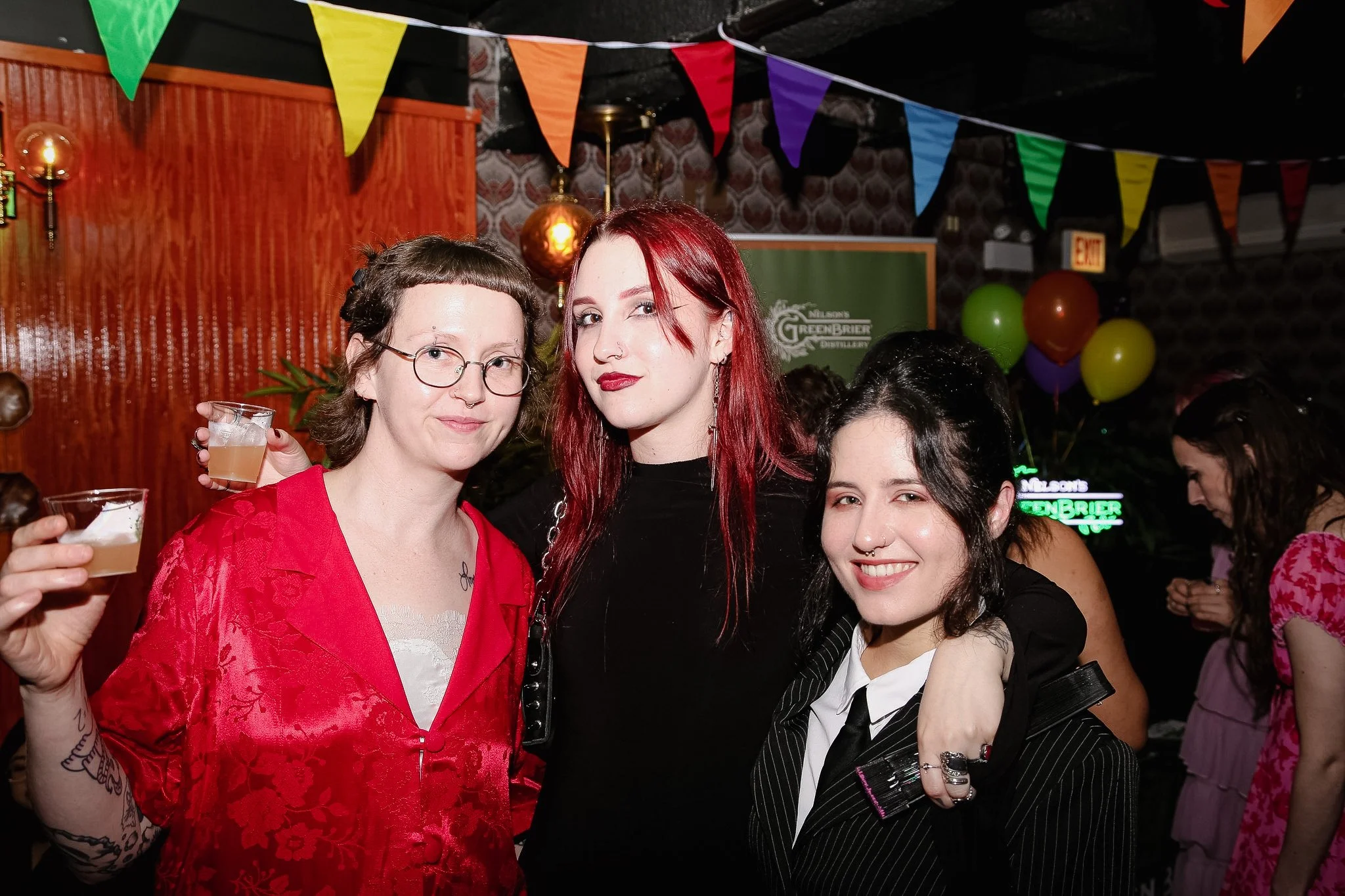 Three queer people stand smiling at the camera. They are at a Queer Prom Event in a lesbian bar in Chicago. Pride decorations hang behind them.