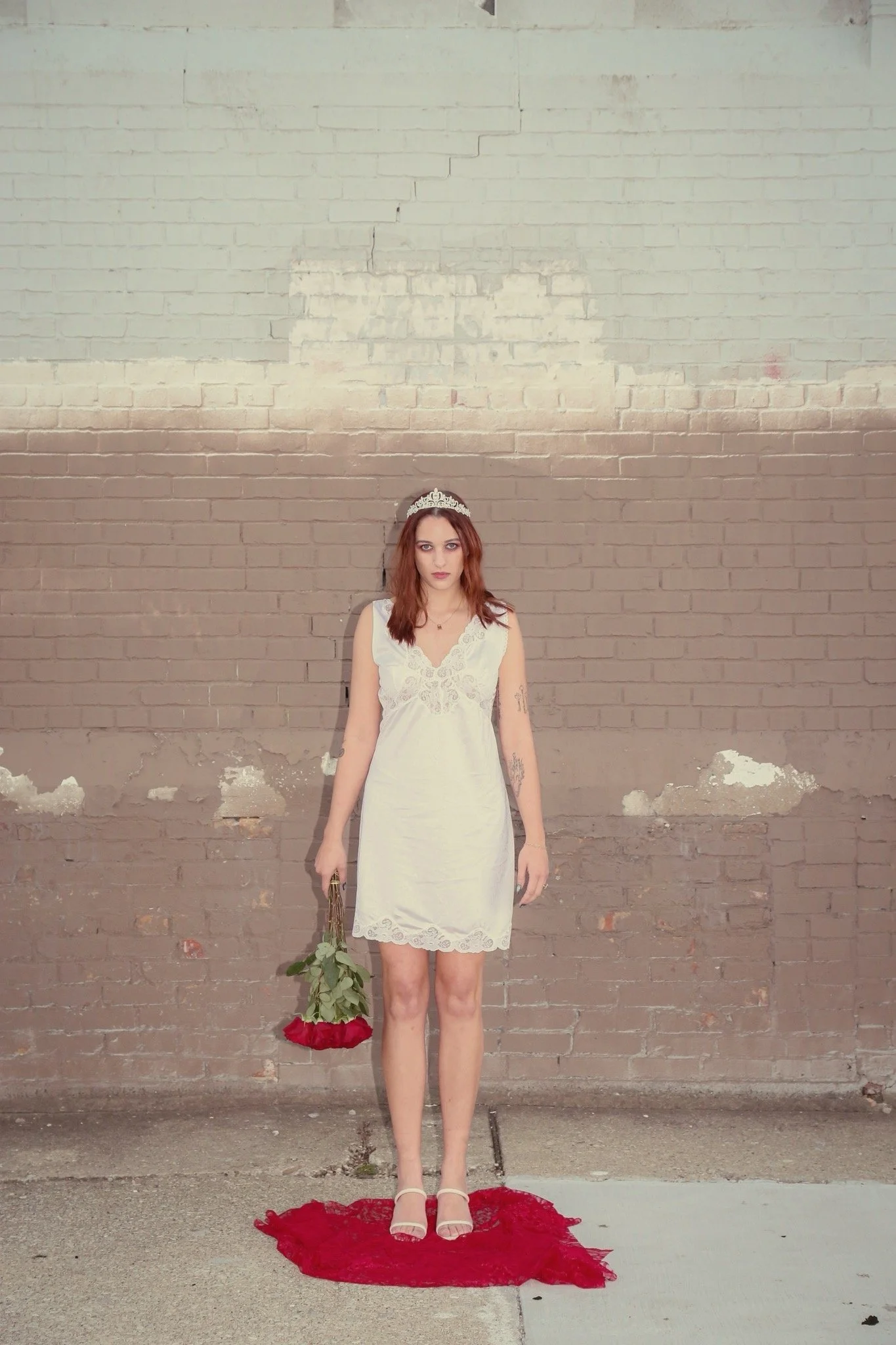 A woman in a white silk dress with a silver crown on her head is holding red roses by her side. She stands in front of a brick wall on top of a red blanket. 