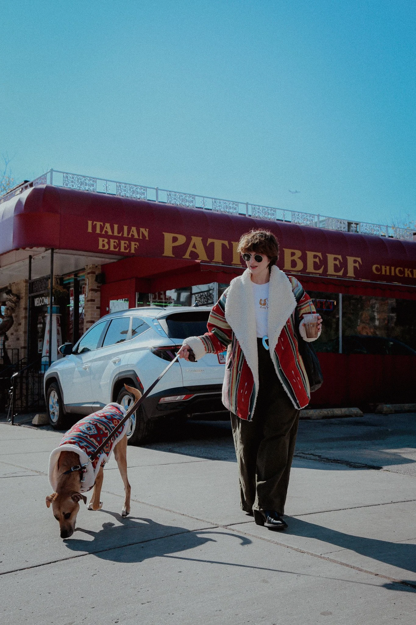 A woman walks her dog in front of an Italian beef restaurant. The person has short brown hair and is wearing an oversized winter red & white jacket. Her dog has on a matching jacket. 