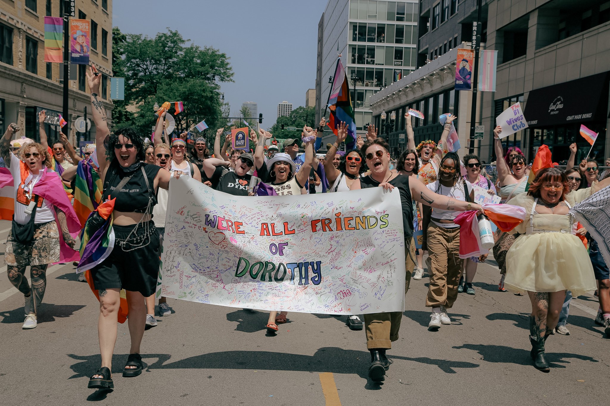 A group of Lesbians march in the Pride Parade holding a sign that says "We're All Friends of Dorothy". They are marching and screaming with joy. 