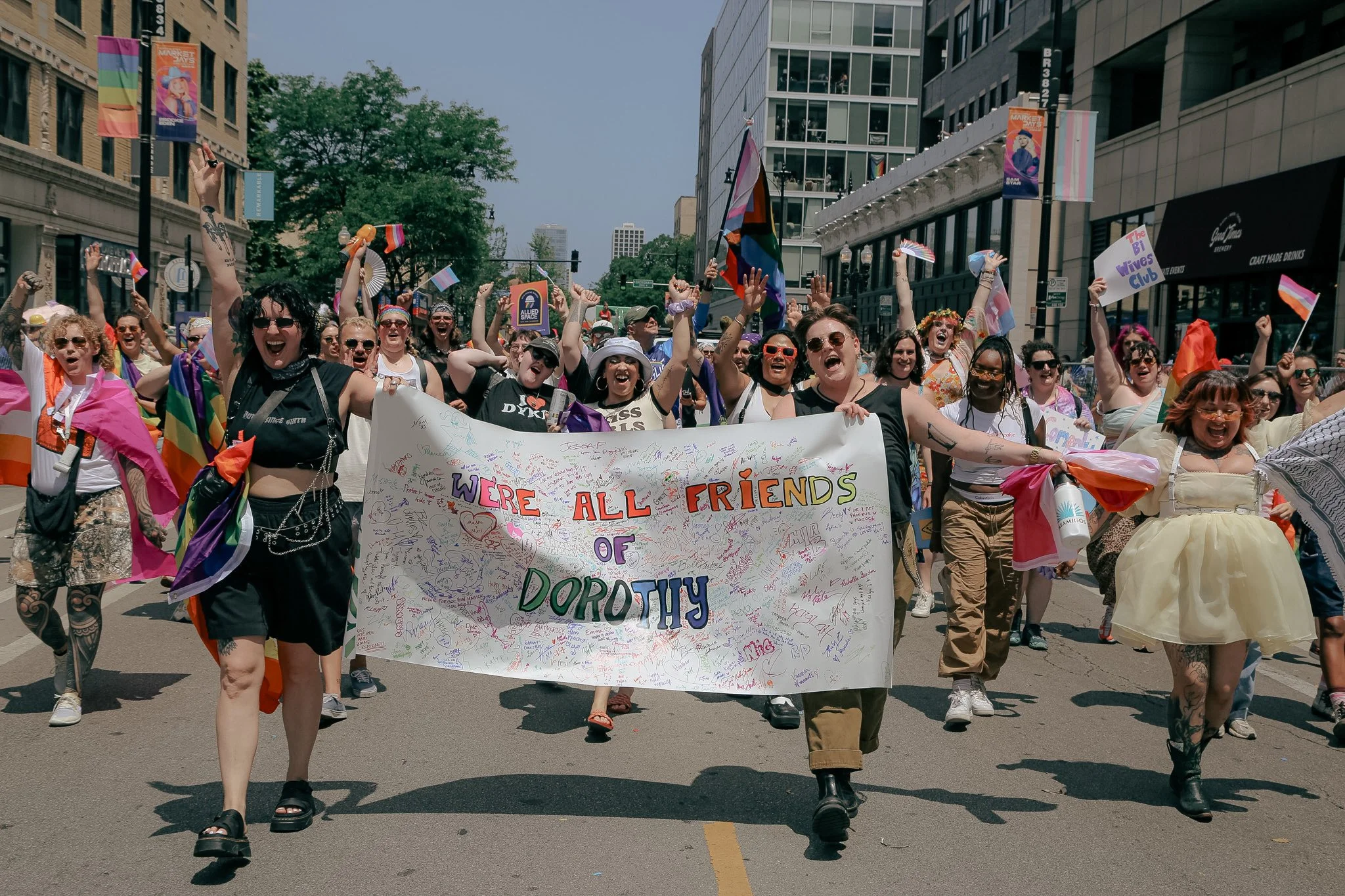 A group of lesbians in the Chicago Pride Parade hold a white sign that says "We're All Friends Of Dorothy". They are marching across the streets of Chicago with joy and pride!