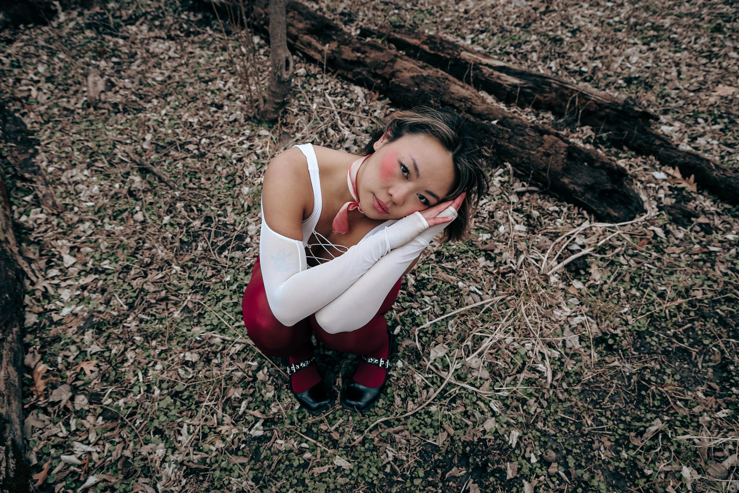A woman in a red top, red tights and white gloves sits in a field in the forest looking into the camera with her hands on her face and a serious expression.