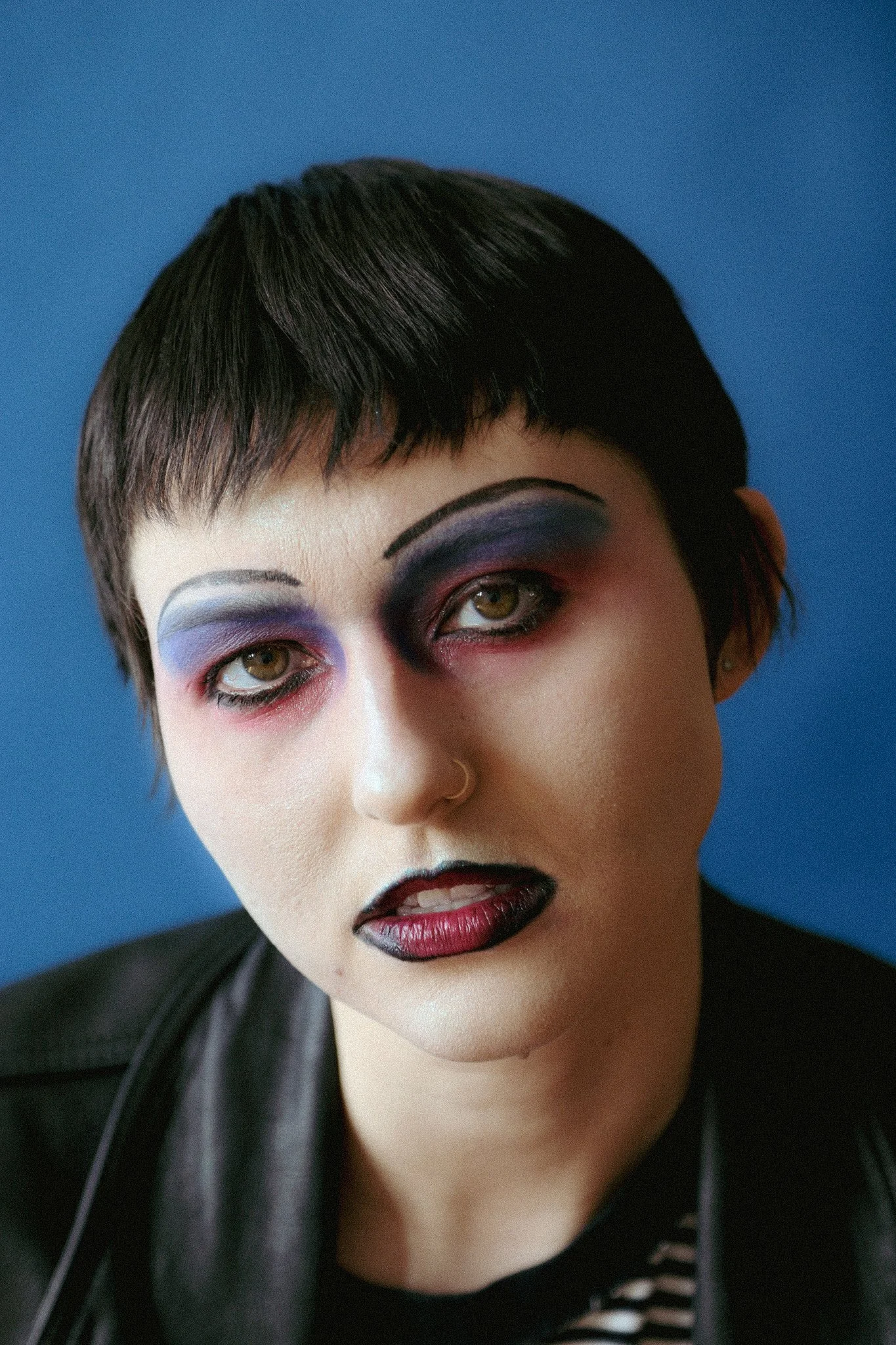 A queer person with blue, pink & black makeup looks seriously into the camera with a blue background. They have on a black leather jacket.
