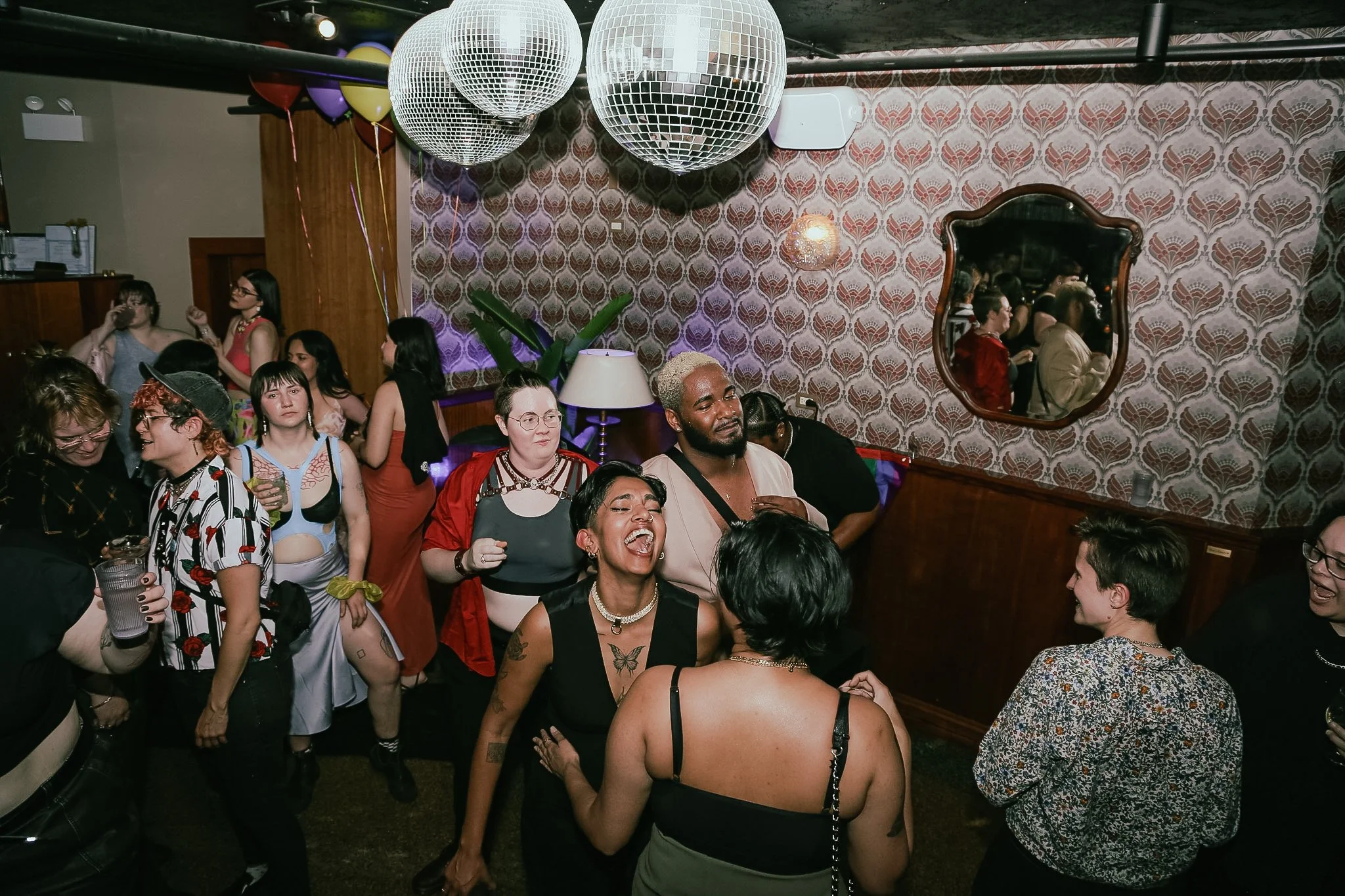 A group of queer individuals are dancing and screaming in a lesbian bar in Chicago under a disco ball. A DJ spins in the corner in front of a giant mirror.