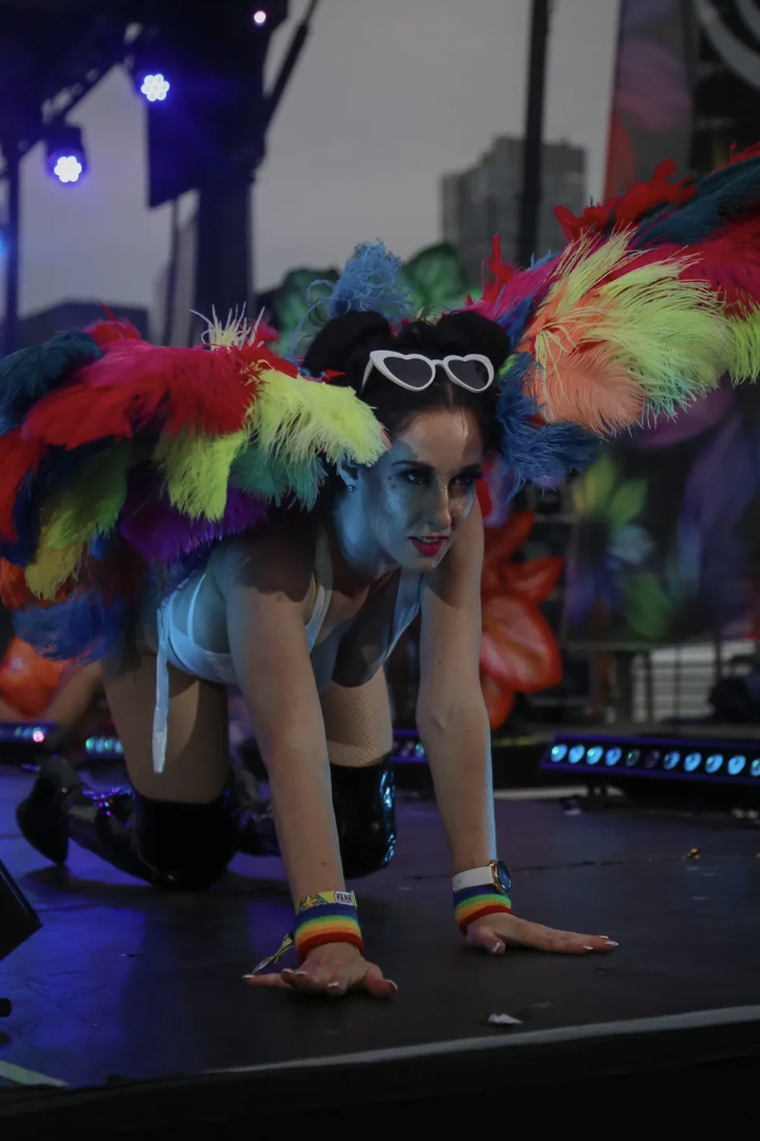A queer person crawls on a stage wearing rainbow feathers and white-hear shaped sunglasses. She has brown hair and glittery makeup. She looked into the crowd with an excited look. 