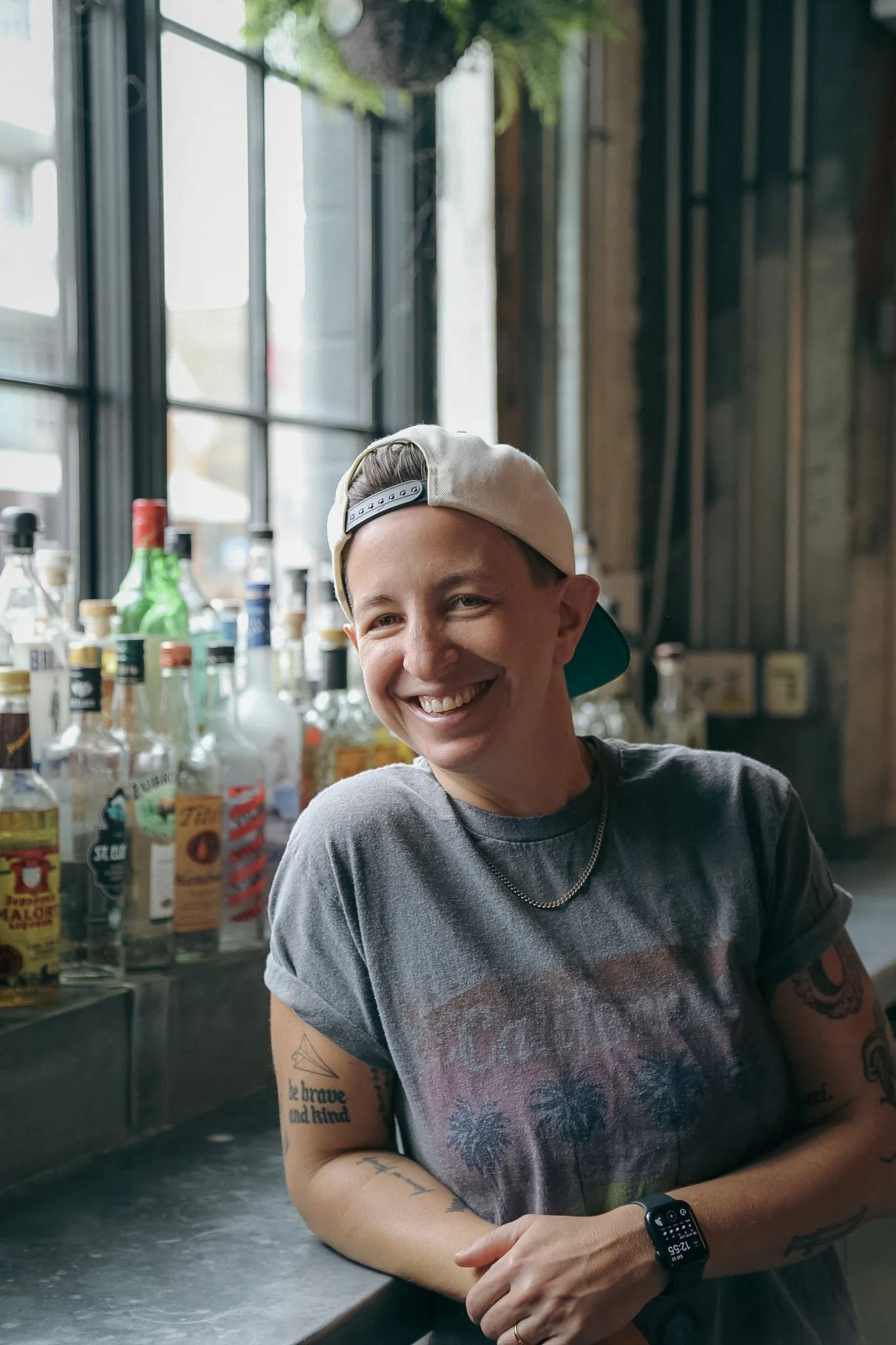 A queer person with a white backwards hat and a grey t-shirt is smiling while leaning on a metal bar. Alcohol bottles lean against a window with greenery above their head.