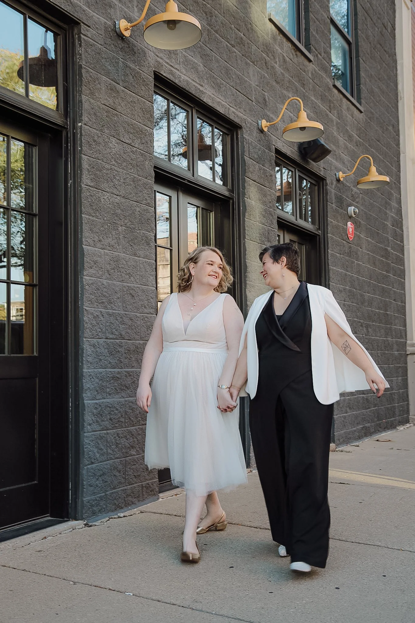 Two queer people walk hand in hand in front of a black brick building. They have just gotten married and are looking into each other's eyes laughing and holding hands. One person wears an black and white pantsuit while the other wears a white dress.