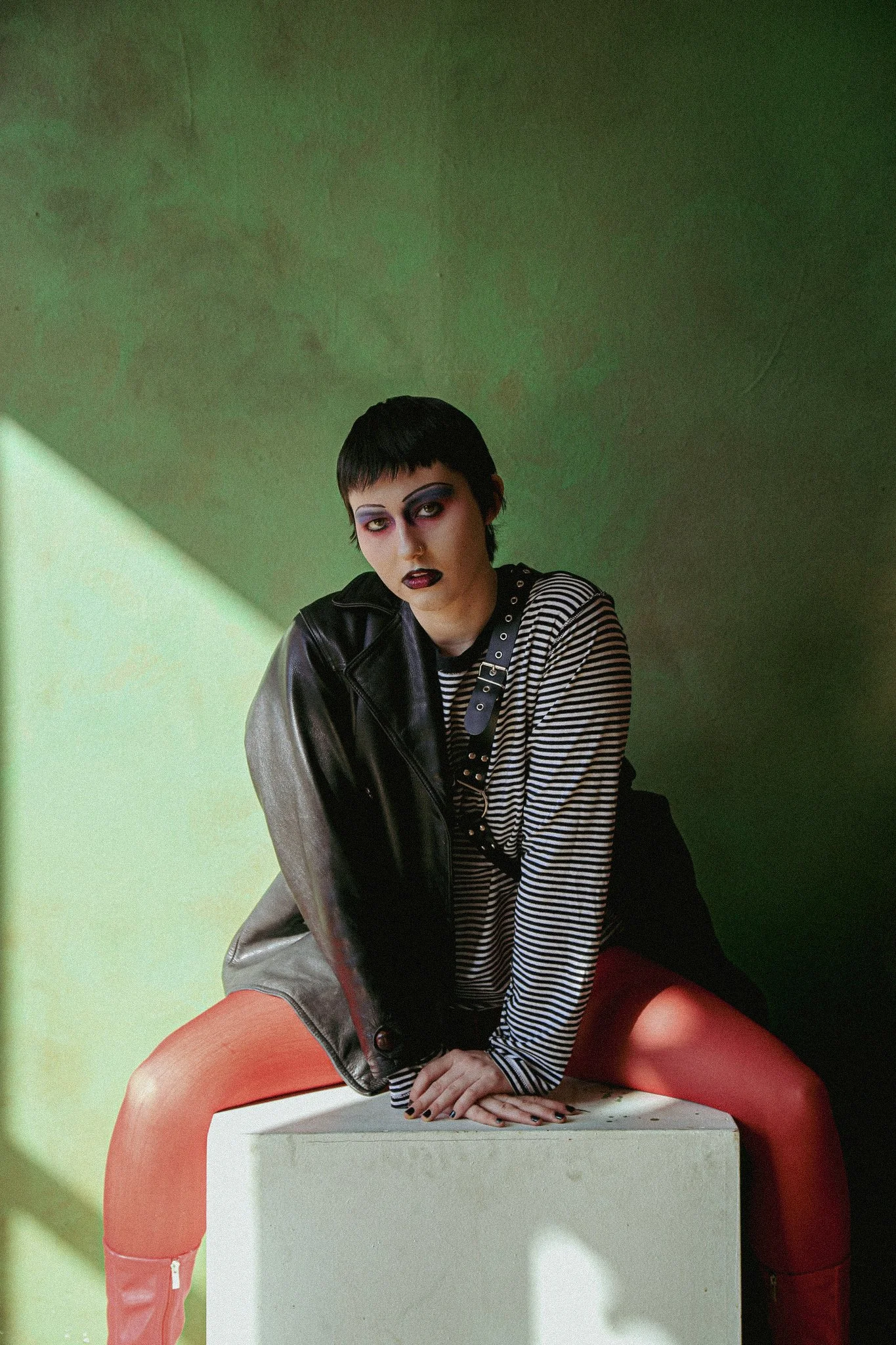 A queer individual sits on a white box wearing red rights, leather jacket and a striped black & white shirt. They have black short hair and are wearing goth-like makeup. They are sitting in front of a green background.