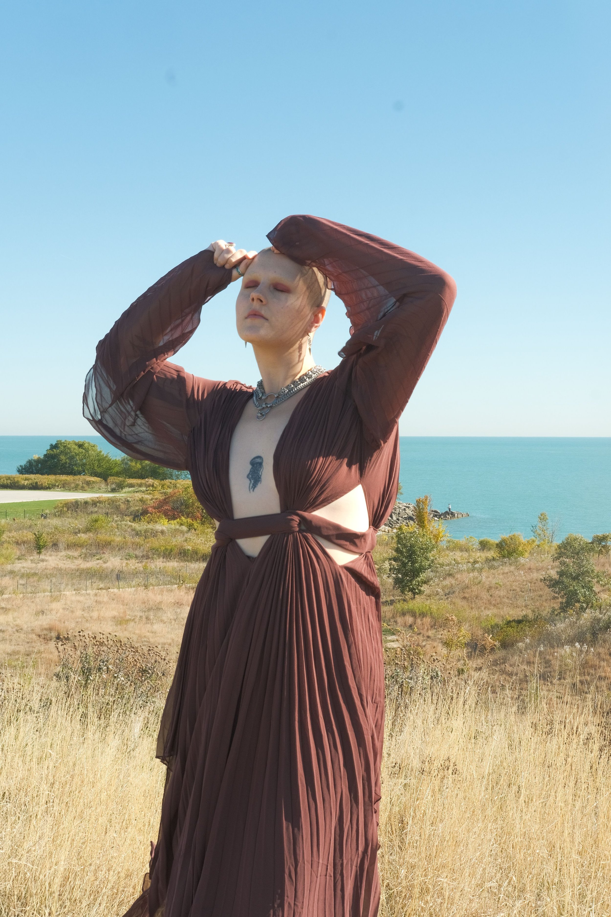 A lesbian stands in a field of green grass with the water behind her in the distance. She has on a brown cutout dress, silver jewelry and her hands are atop her head. She has her eyes closed.