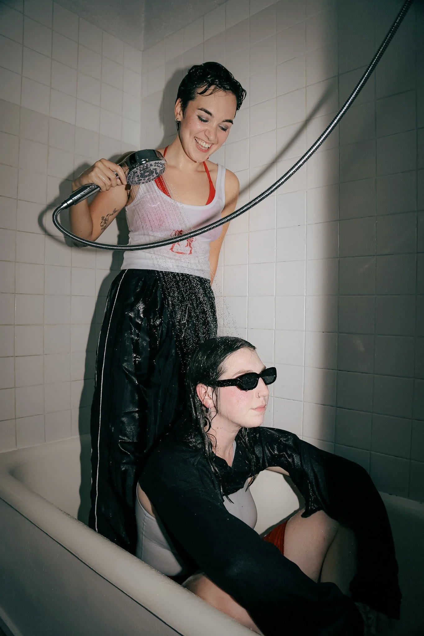 Two queer people that are fully clothed are in a bathtub. One person stands in the shower with the shower head over top of another person who is sitting in the bathtub getting soaked by the water. Both are wearing black, white and red clothing.