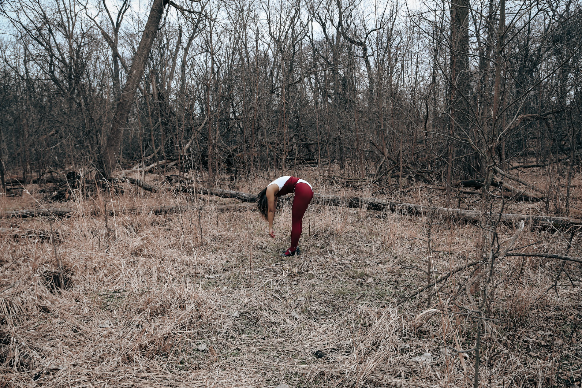 A woman strand bent over with her arms handing in the barren woods. She has on red tights with a white and red top. Her face is hidden by her hanging arms.