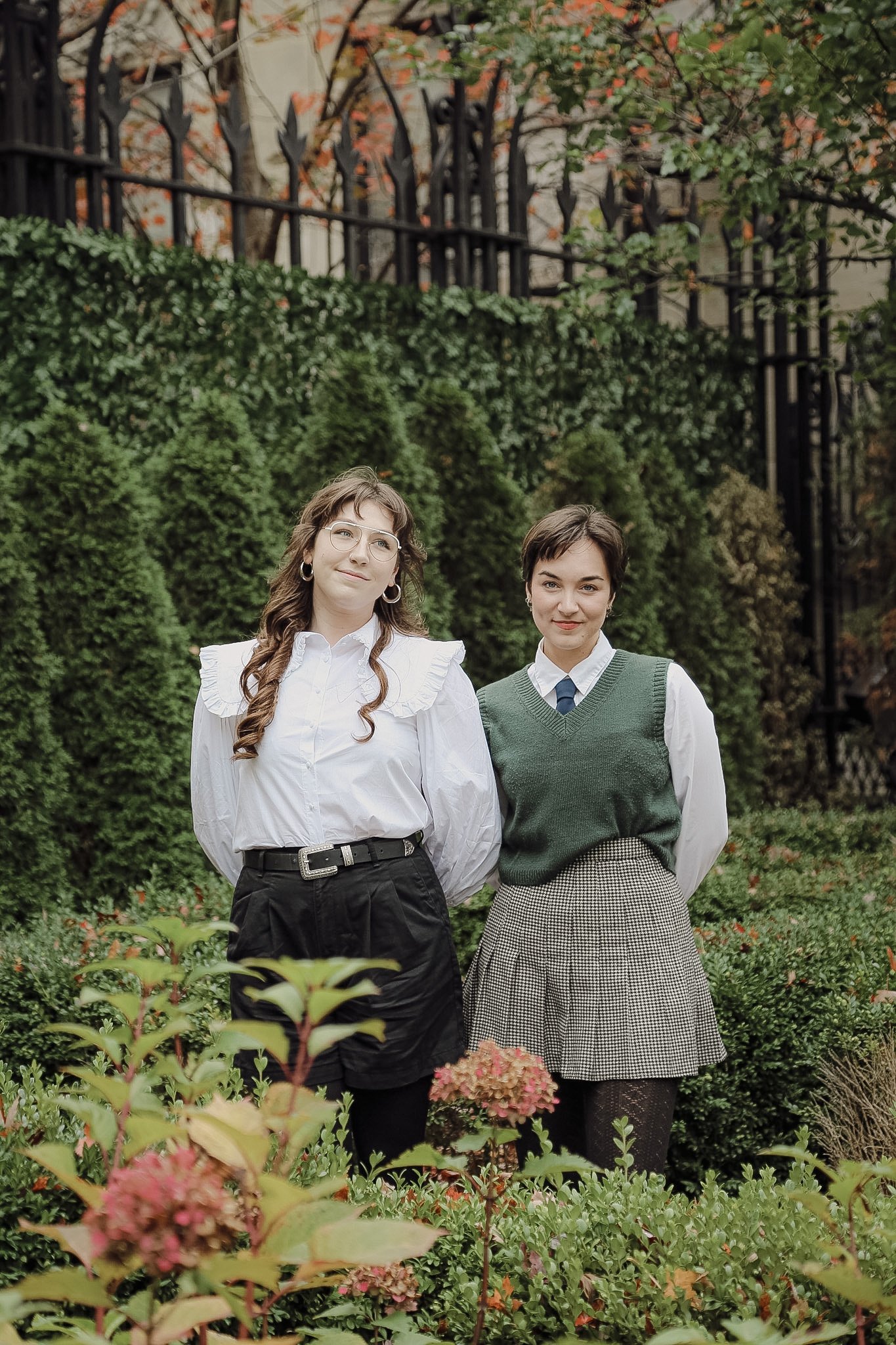 Two lesbians stand side by side in a whimsical garden. They are wearing 90's inspired outfits and looking content.