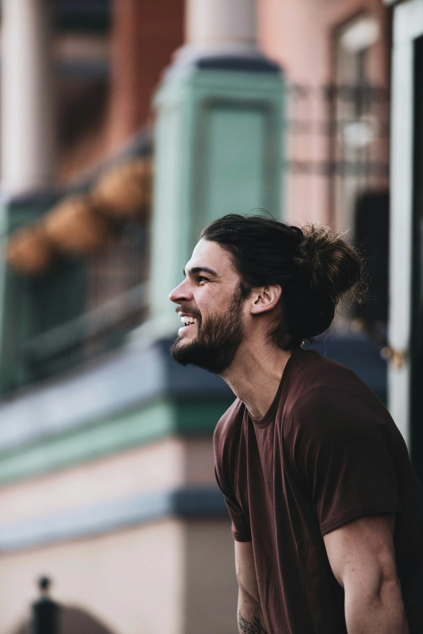 A man with long black hair tied in a bun, beard, and tattoos, smiling and looking away, wearing a brown t-shirt, in an urban setting with colorful buildings and a green trash can in the background.