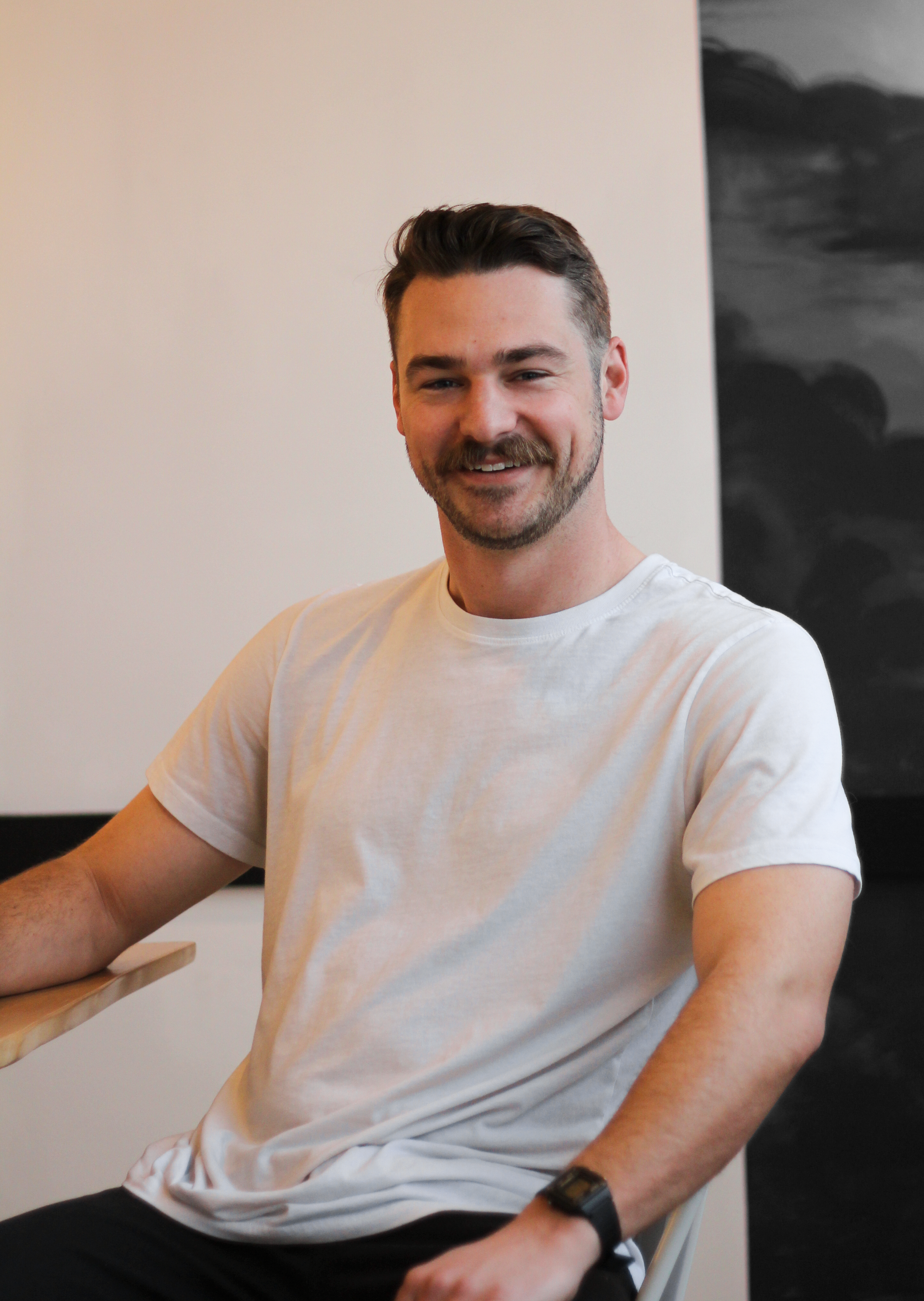 A smiling man with dark hair and a beard, wearing a white T-shirt, sitting at a table indoors with a black and white painting in the background.