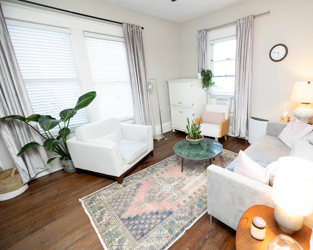 Bright living room with large windows, white couches, a green coffee table, plants, a patterned rug, and light-colored curtains.
