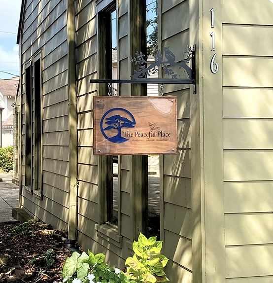 A wooden sign reading 'The Peaceful Place' hanging outside a building with green siding and the street number 116, decorated with a tree and butterfly design.