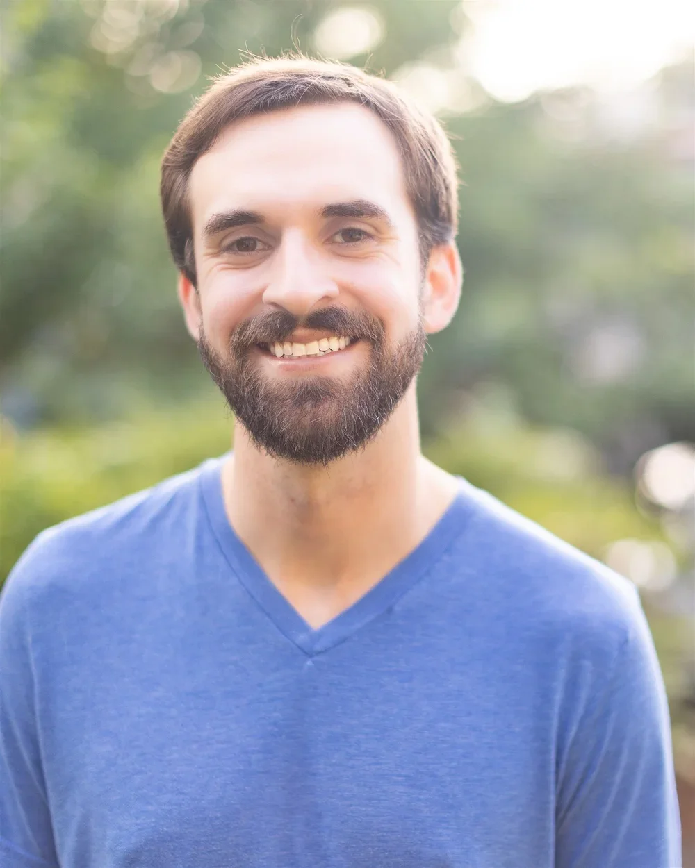 A smiling man with a beard and brown hair, wearing a blue V-neck shirt, standing outdoors with blurred greenery in the background.