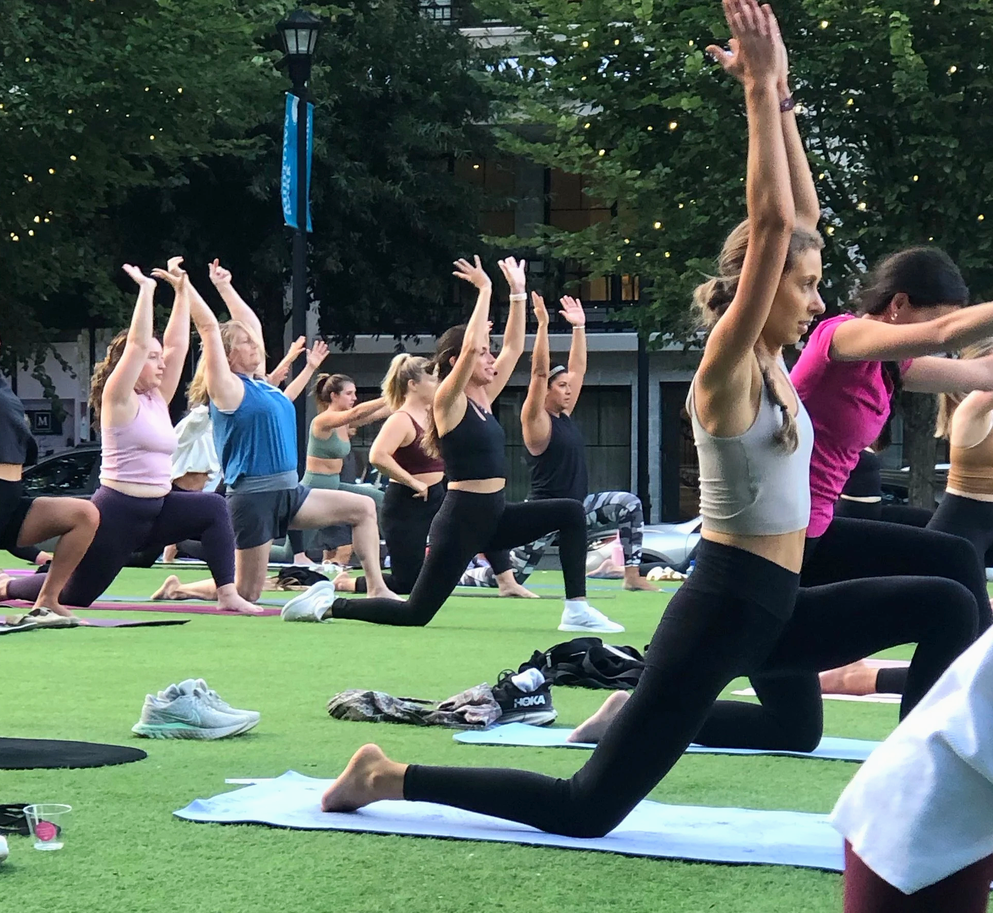 A group of people participating in an outdoor yoga class on a grassy surface in the evening, with trees and a lamppost in the background, all in a kneeling pose with arms raised overhead.