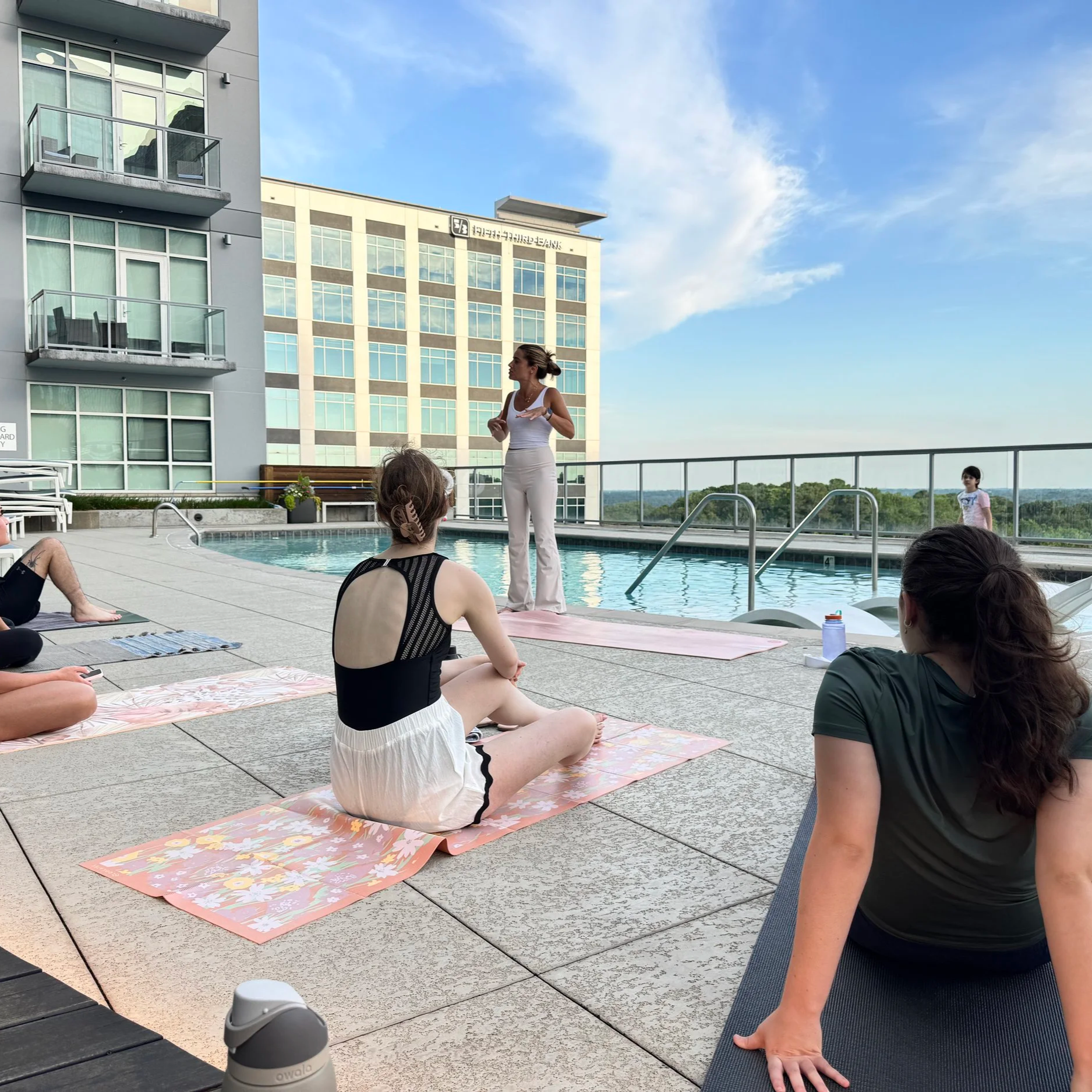 A yoga instructor leading a class on a rooftop pool deck during daytime. Participants are seated on yoga mats in a cross-legged position, facing the instructor. The pool is behind them, and the sky is partly cloudy.