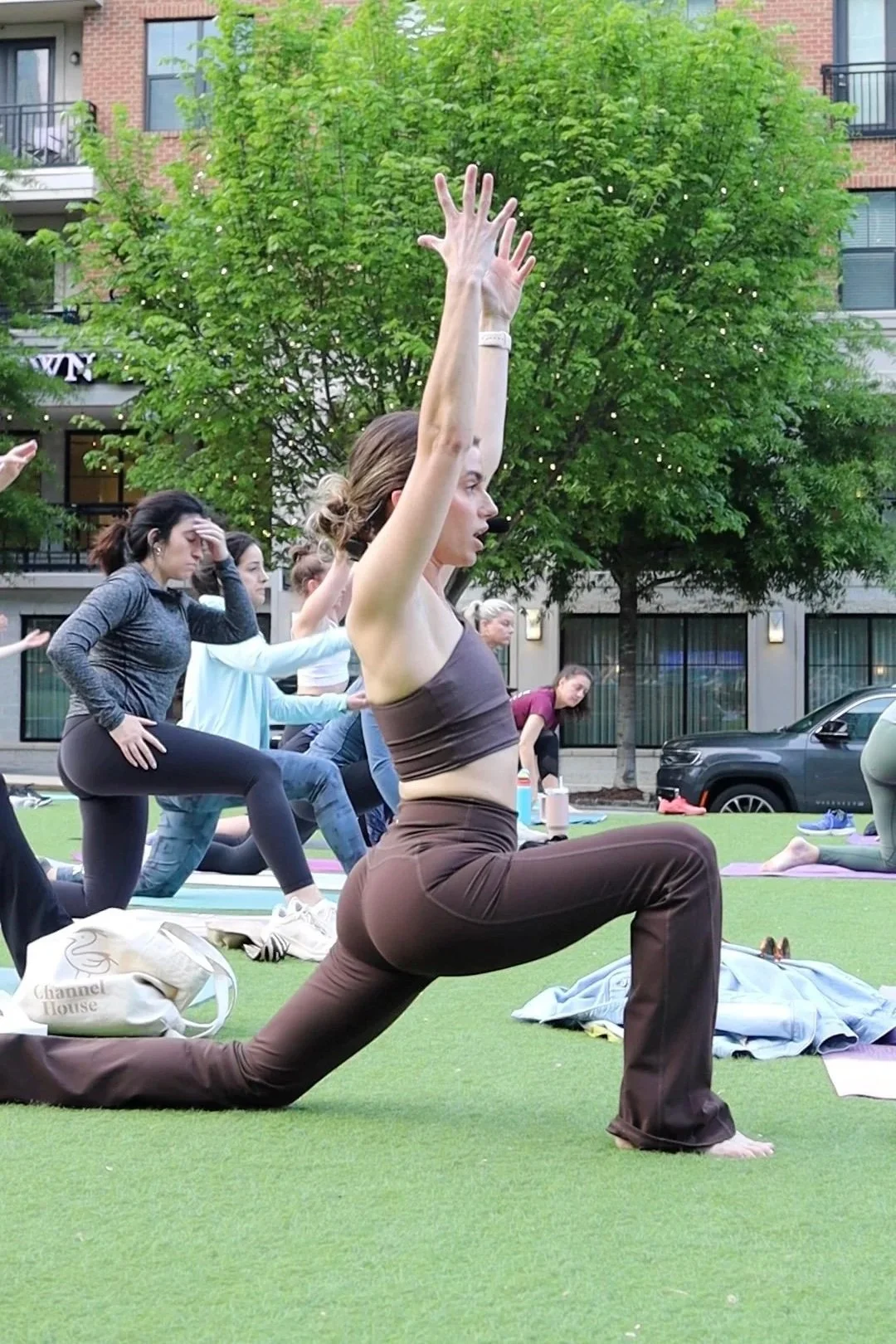Group of people practicing yoga outdoors on a grassy area, with a woman in the foreground performing a lunge pose with arms raised above her head, surrounded by trees and buildings.