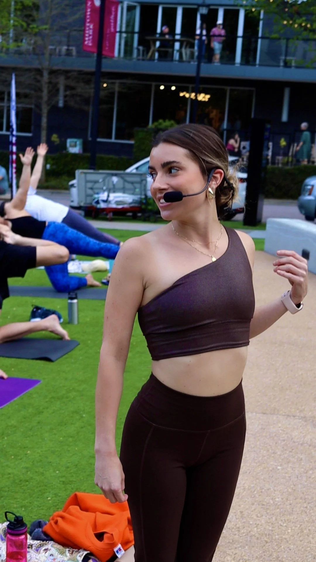 A young woman with a microphone headset leading a yoga class outdoors, with other participants lying on mats and stretching in the background.