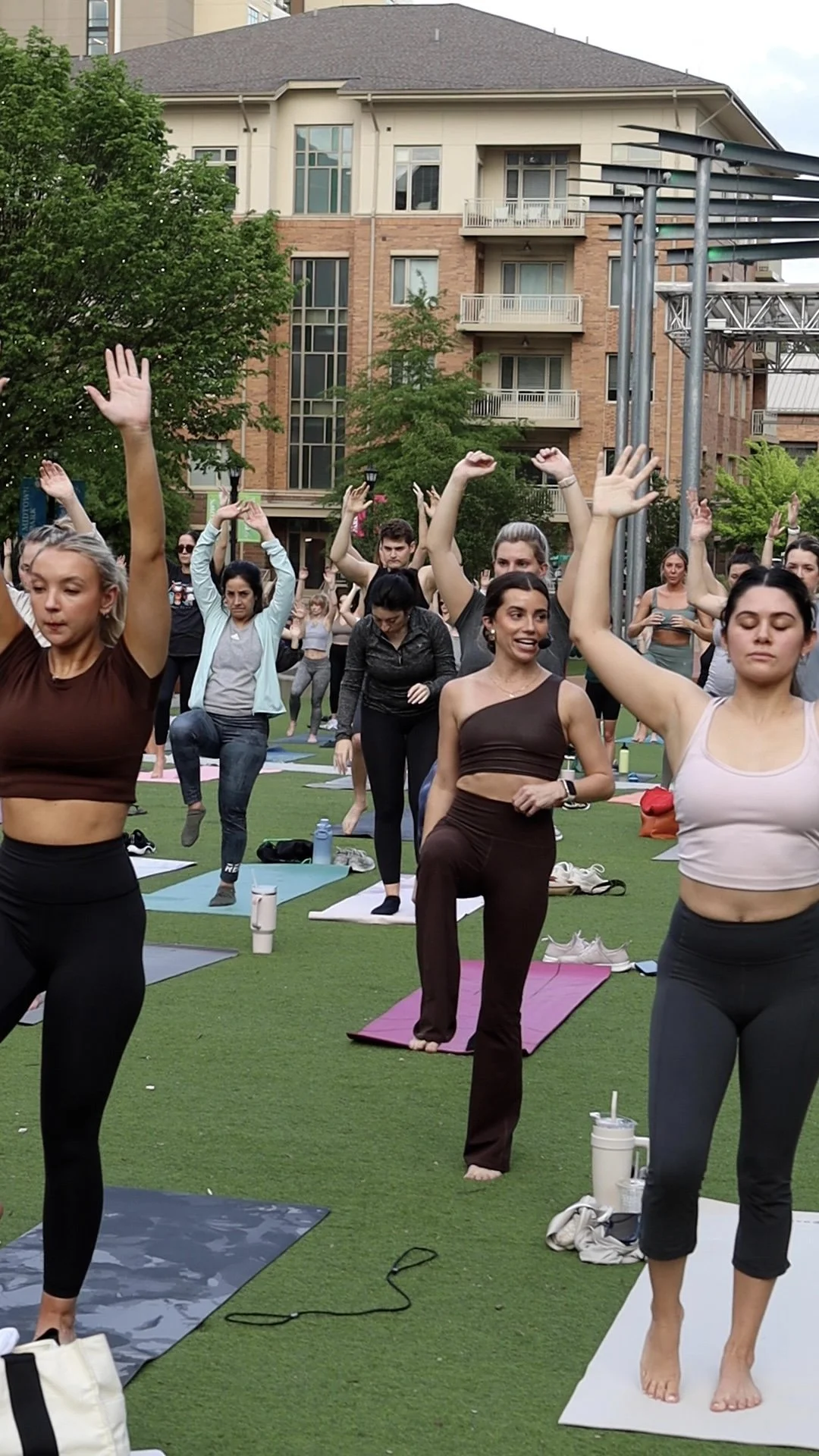 A group of women participating in an outdoor yoga or fitness class on green artificial turf, with trees and a multi-story building in the background.