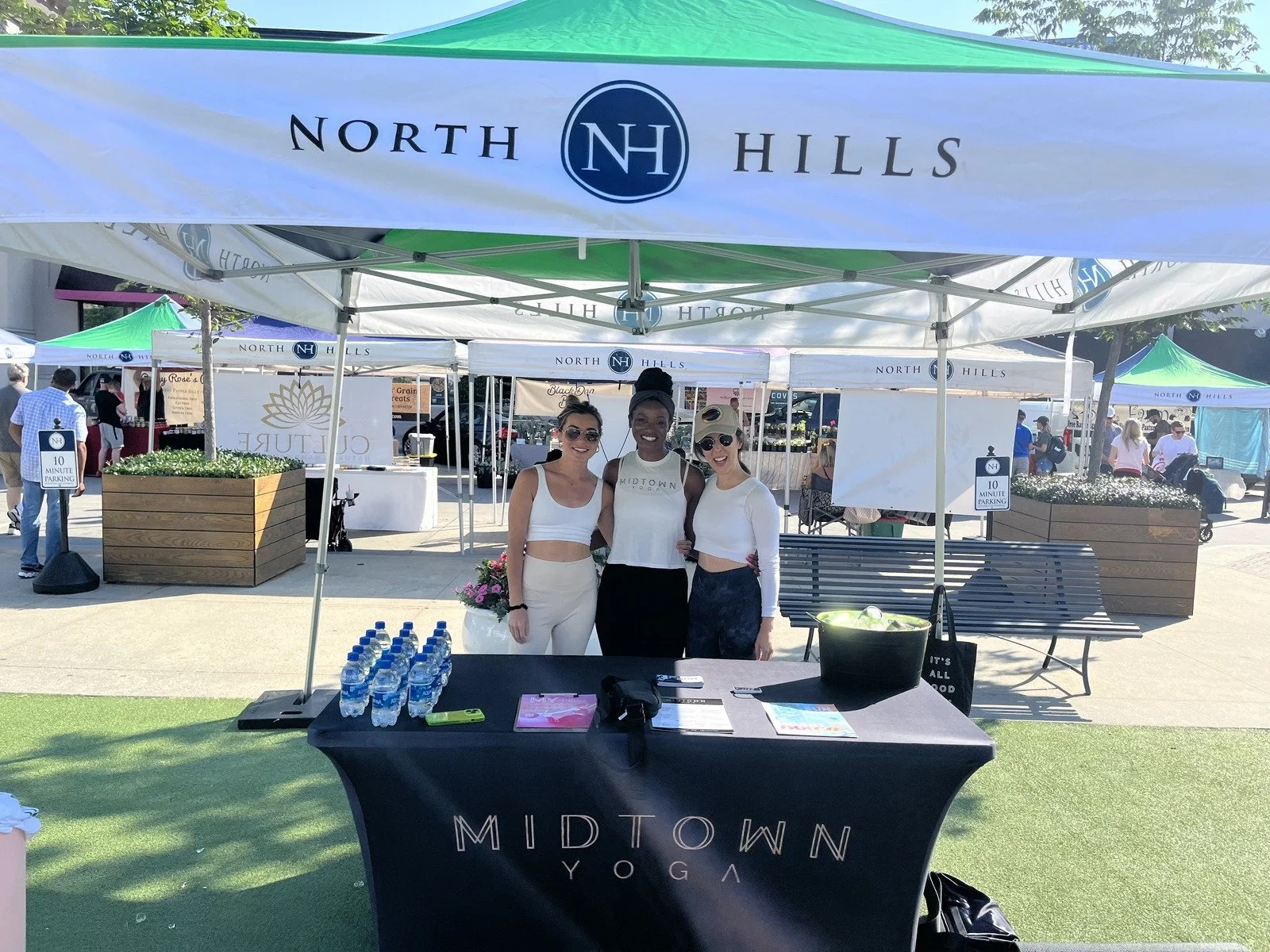 Three women standing behind a table at an outdoor event under a tent labeled 'North Hills'. The table has bottled water, flyers, and a black bag, with a sign that says 'Midtown Yoga'. In the background, multiple tents and people are visible.