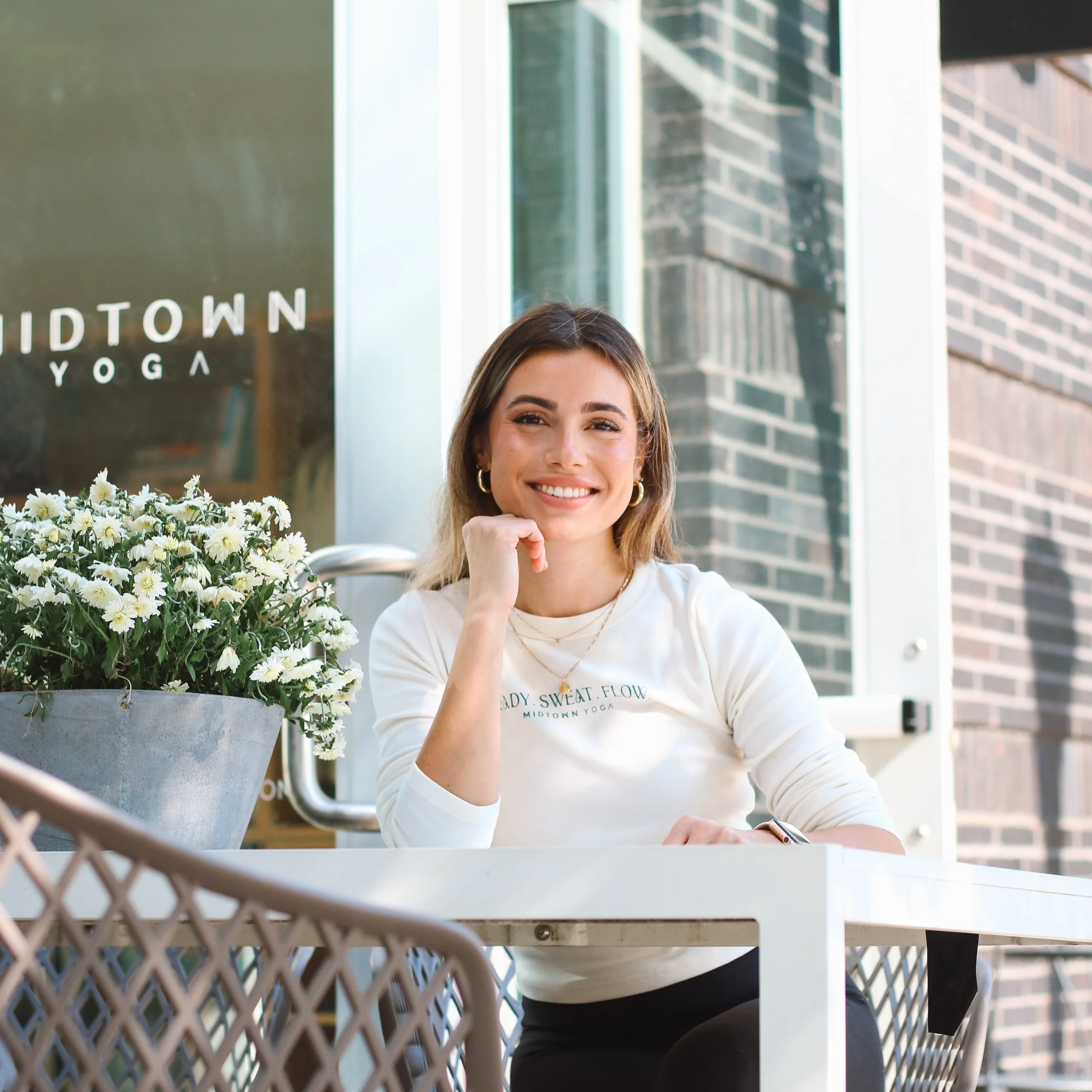 Yoga teacher Daniela Burn sitting at an outdoor table near Midtown Yoga in Raleigh NC, with a large pot of white flowers next to her, in front of a brick building.