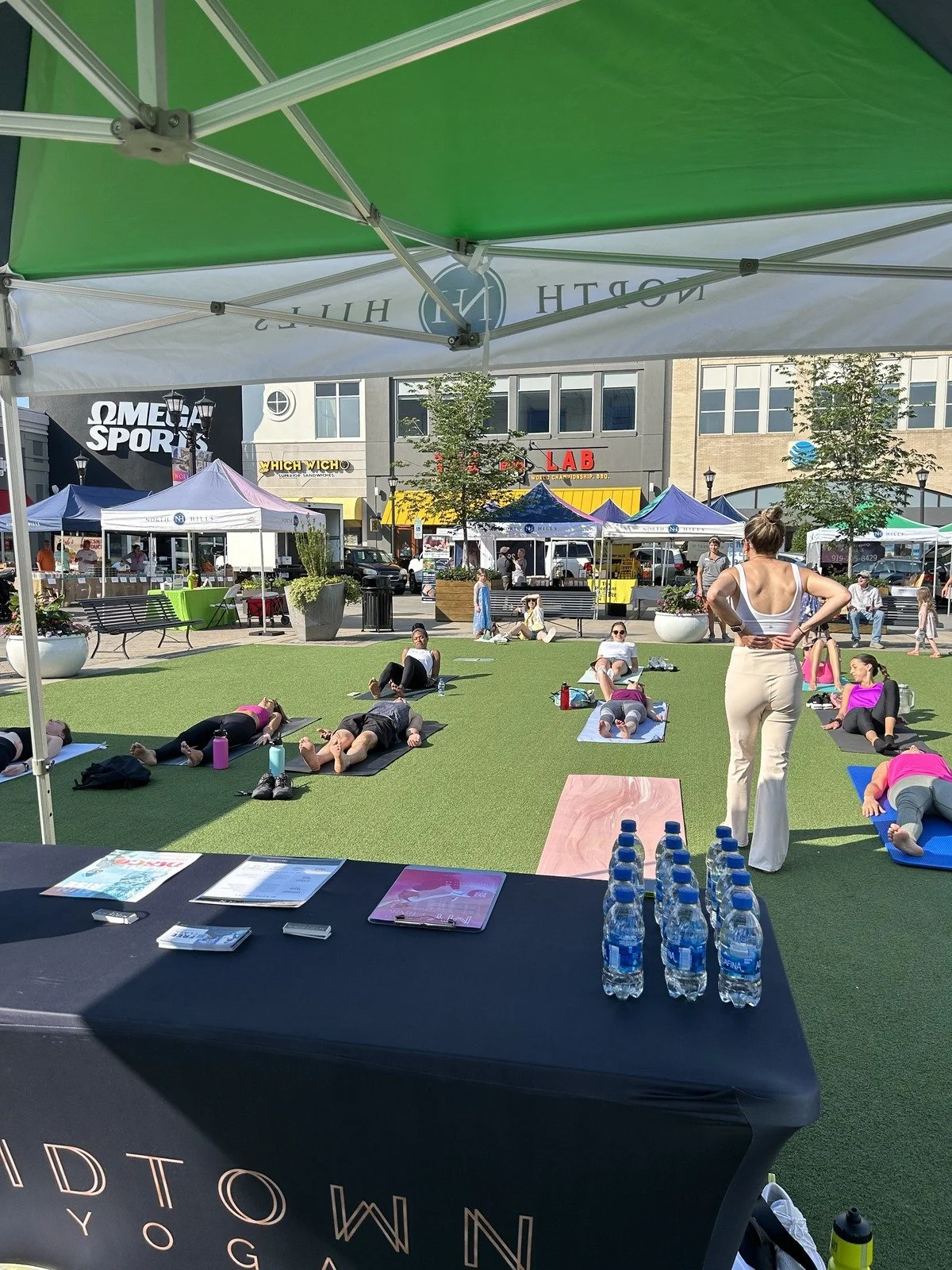 An outdoor yoga class on a city street with participants lying on yoga mats, a yoga instructor standing at the front, and tents and shops in the background.