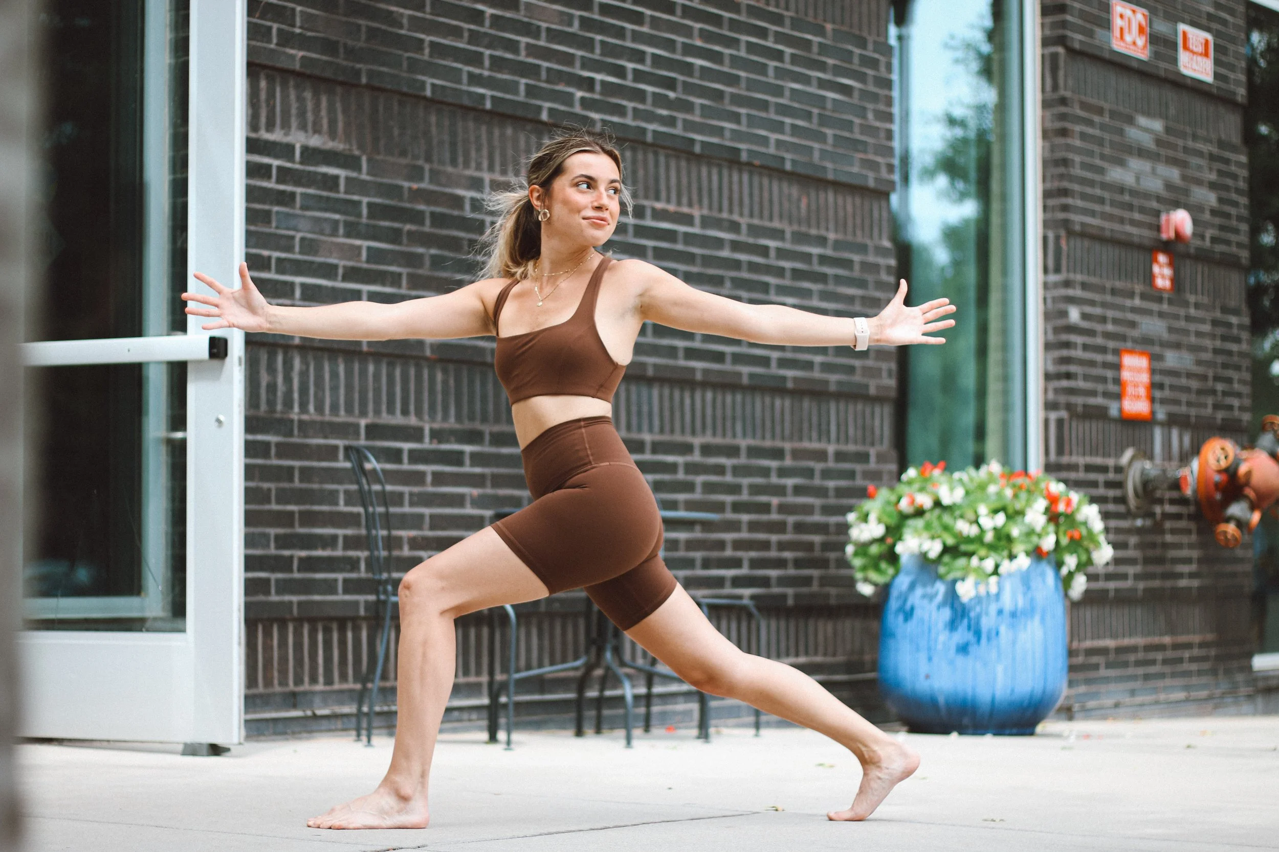 Yoga teacher Daniela Burn in brown workout clothes and a white watch practicing yoga outside near a brick building with a blue flower pot and some benches.