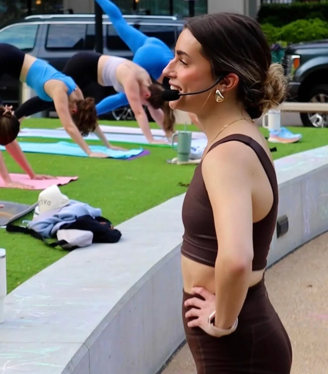 Yoga teacher Daniela Burn with a microphone headset overseeing an outdoor yoga class with participants on yoga mats.
