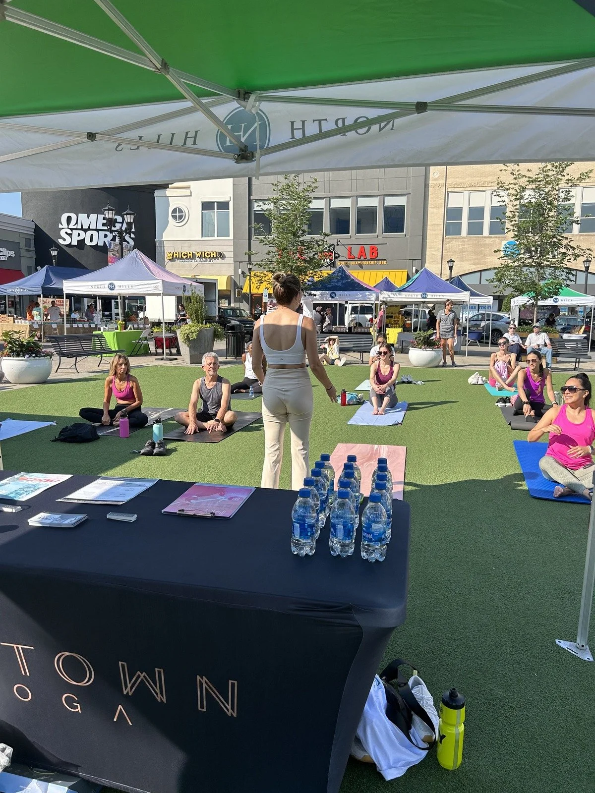 People participating in a yoga class on an outdoor grassy area, with tents and shops in the background.