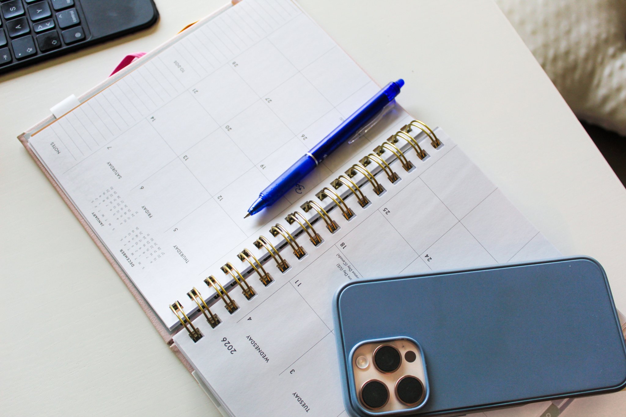 An open spiral-bound calendar on a desk with a blue pen resting on it, a black keyboard partially visible at the top left, and a smartphone with a camera lens on the bottom right