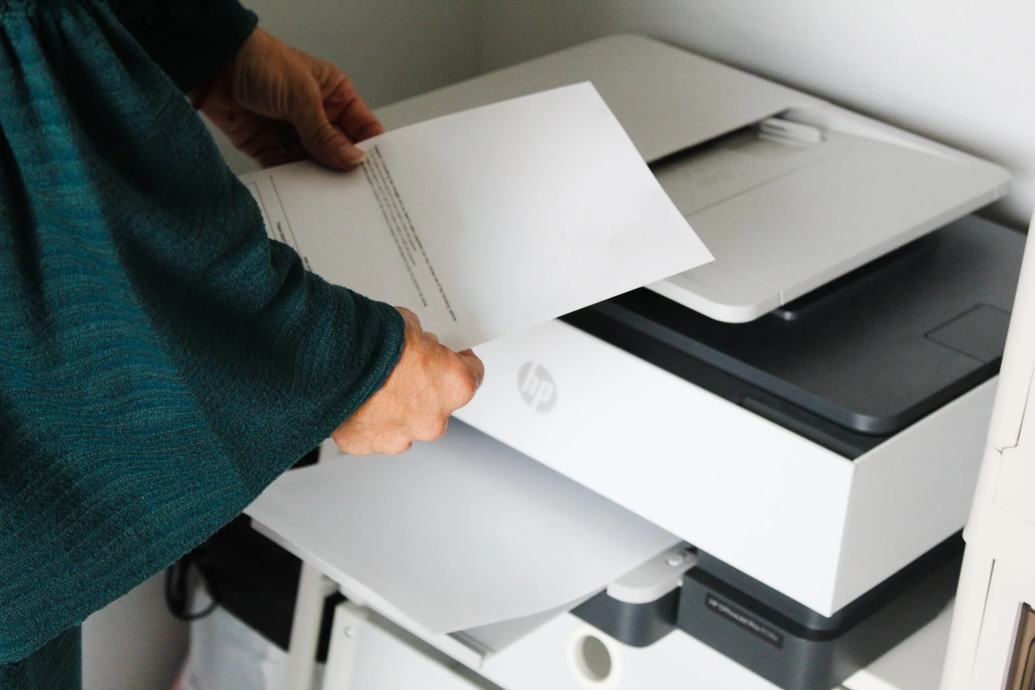 Person holding a white sheet of paper over a white printer with a digital display, situated on a white cart with shelves.