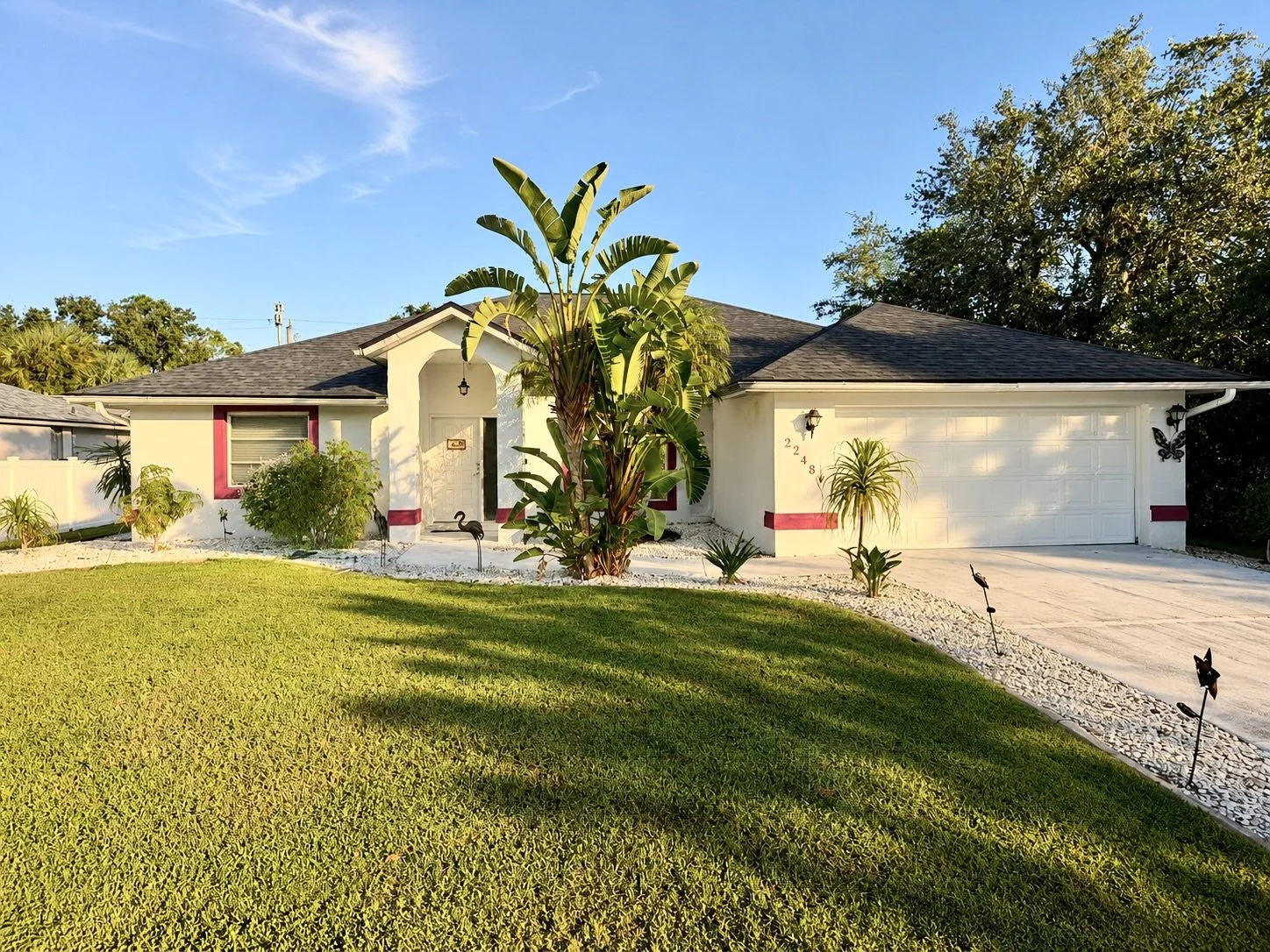 Single-story house with a white exterior, dark shingle roof, and pink trim around the window and door. The front yard features a well-maintained lawn, tropical plants, and decorative black swan garden ornaments. A concrete driveway leads to a two-car garage, and the sky above is blue with a few clouds.