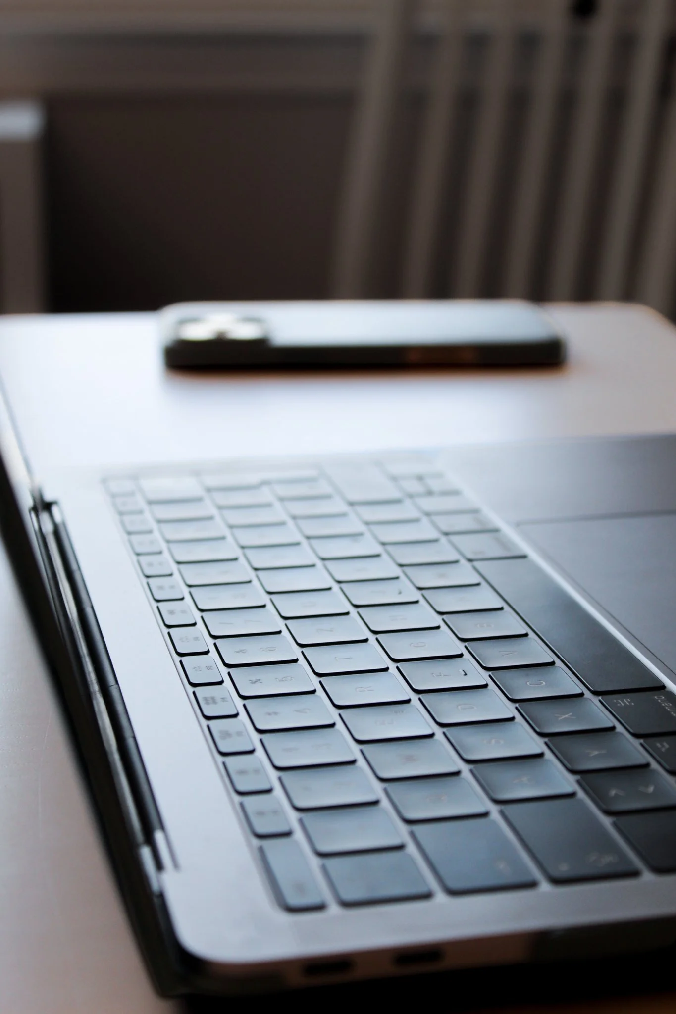 Close-up of a laptop keyboard with a smartphone placed on top, and a blurred background of a chair and furniture.