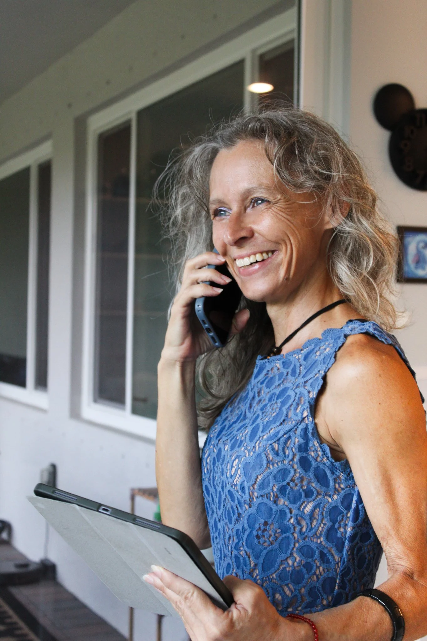 Older woman with curly gray hair smiling while talking on a landline phone indoors, holding a tablet, wearing a blue lace dress and black wristwatch.